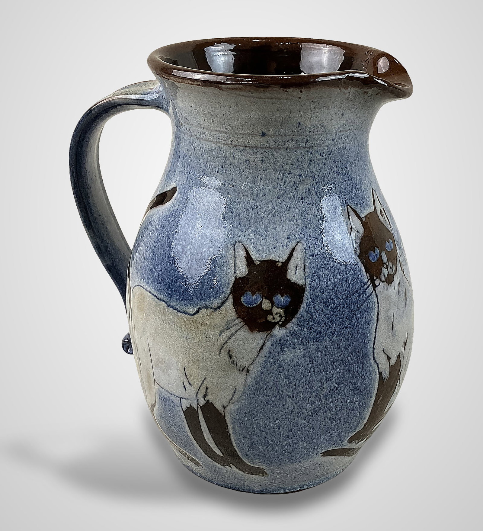 Chelsea Pottery Cat Pitcher: Art pottery piece features a circumference encircled by Siamese cats set against a soft blue ground. The pitcher is signed by Joyce Morgan and includes a Chelsea label affixed to the base. It