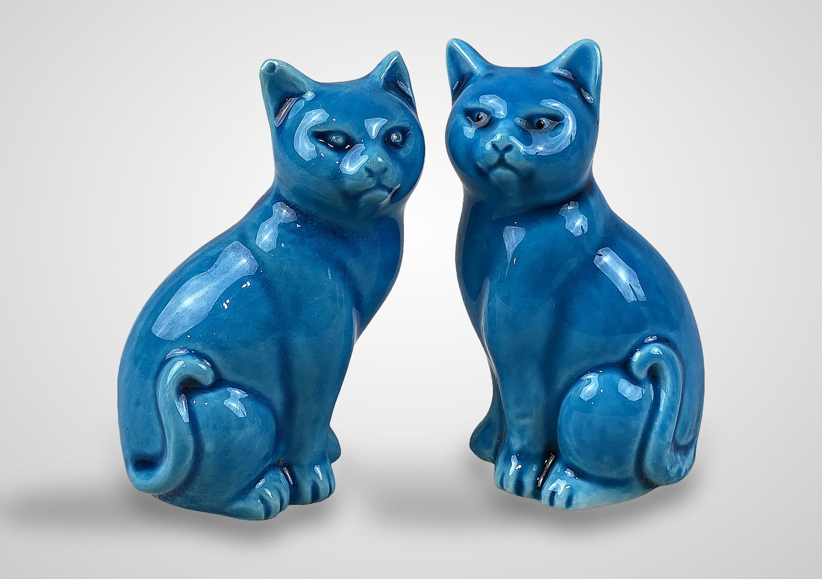 Pair Blue Glazed Porcelain Cats: Charming pair of cats features a rich, vibrant blue glaze and are captured in a seated pose with a sense of naturalism. Each piece measures approximately 3 1/4 in. H x 2 in. W x 1 1/4 in. D. Both are 