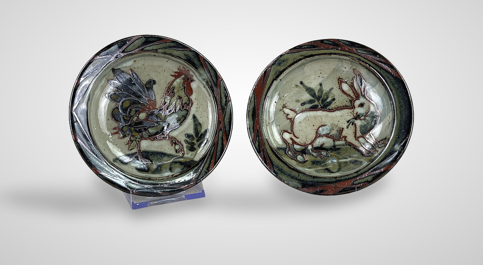 Studio Art Pottery Rooster and Rabbit Bowls: Two handcrafted pottery bowls featuring a speckled glaze. One bowl showcases a rooster in the center painted in shades of brown, green, and orange, with red overlapping lines decorating the rim and a 