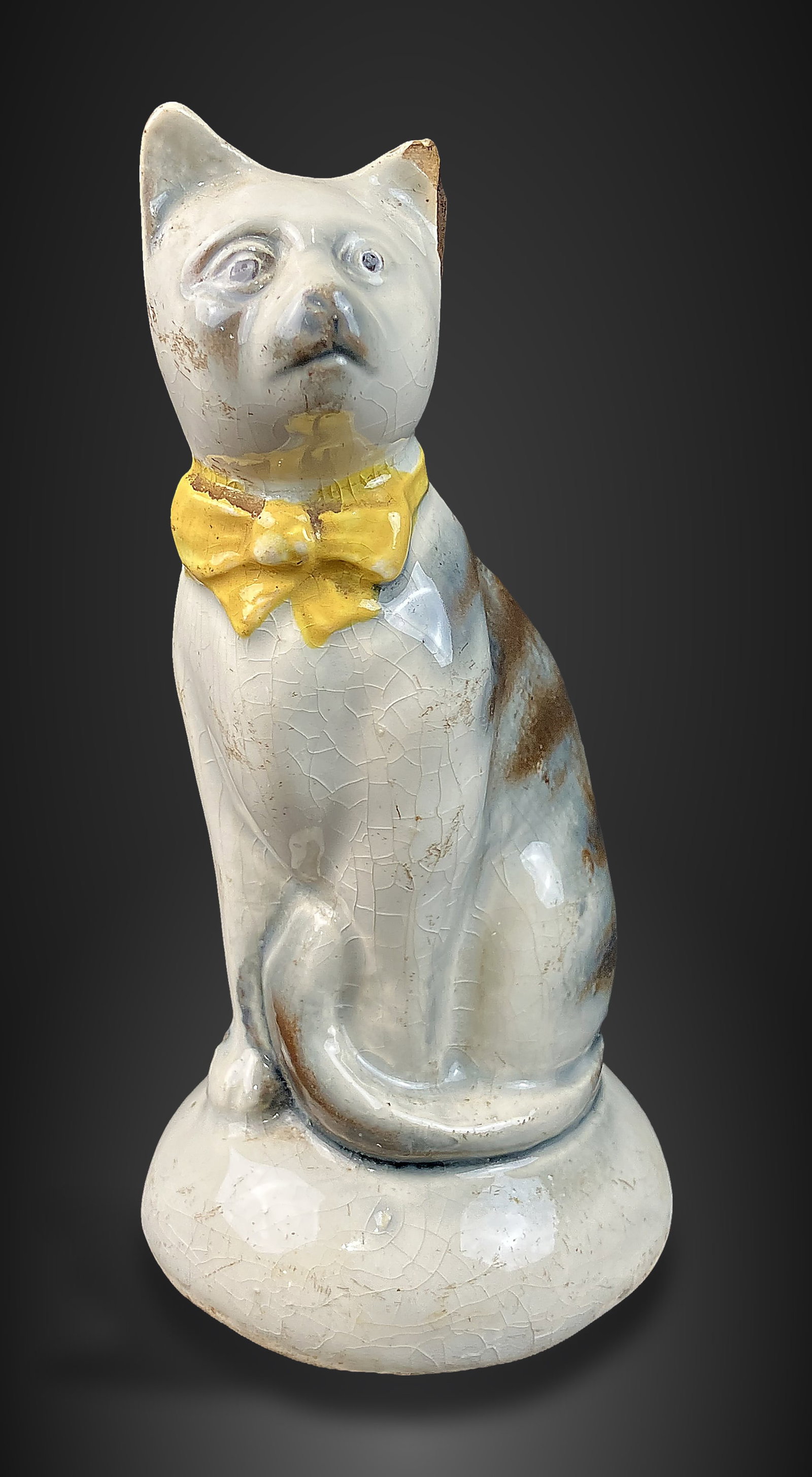 Staffordshire Cat Bank: Elegantly sculpted ceramic bank features a cat detailed with a yellow bowtie and a white face. It measures approximately 4 3/4 in. H x 2 3/8 in. in diameter. The piece shows general wear consistent