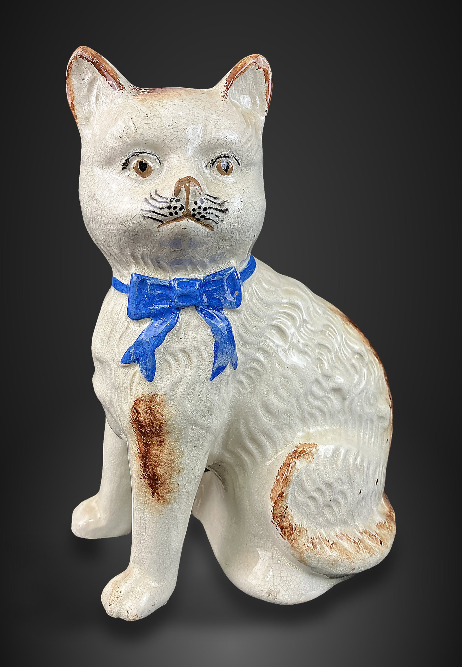 Staffordshire Cat Figurine: Charming figurine depicts a cat in an upright posture, featuring well-defined molded fur and distinctive hand-painted facial features, accented by a blue bow on its collar. It measures approximately