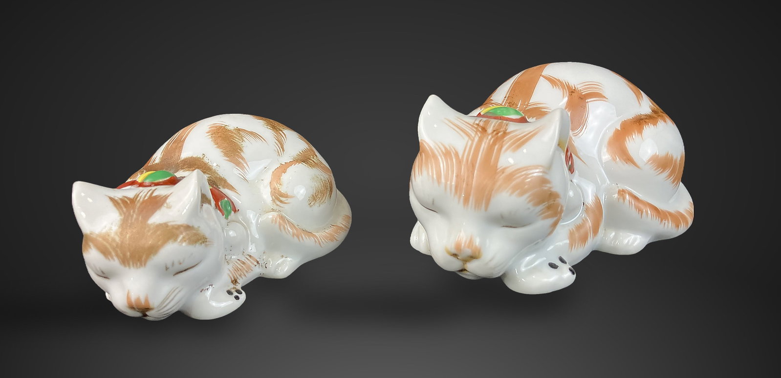 Pair of Japanese Kutani Sleeping Cats: Pair of hand-painted porcelain figurines depicts cats in resting poses. They are decorated with ginger-toned stripes and delicate gilt detailing, accented by vibrant red, green, and yellow collars. Th