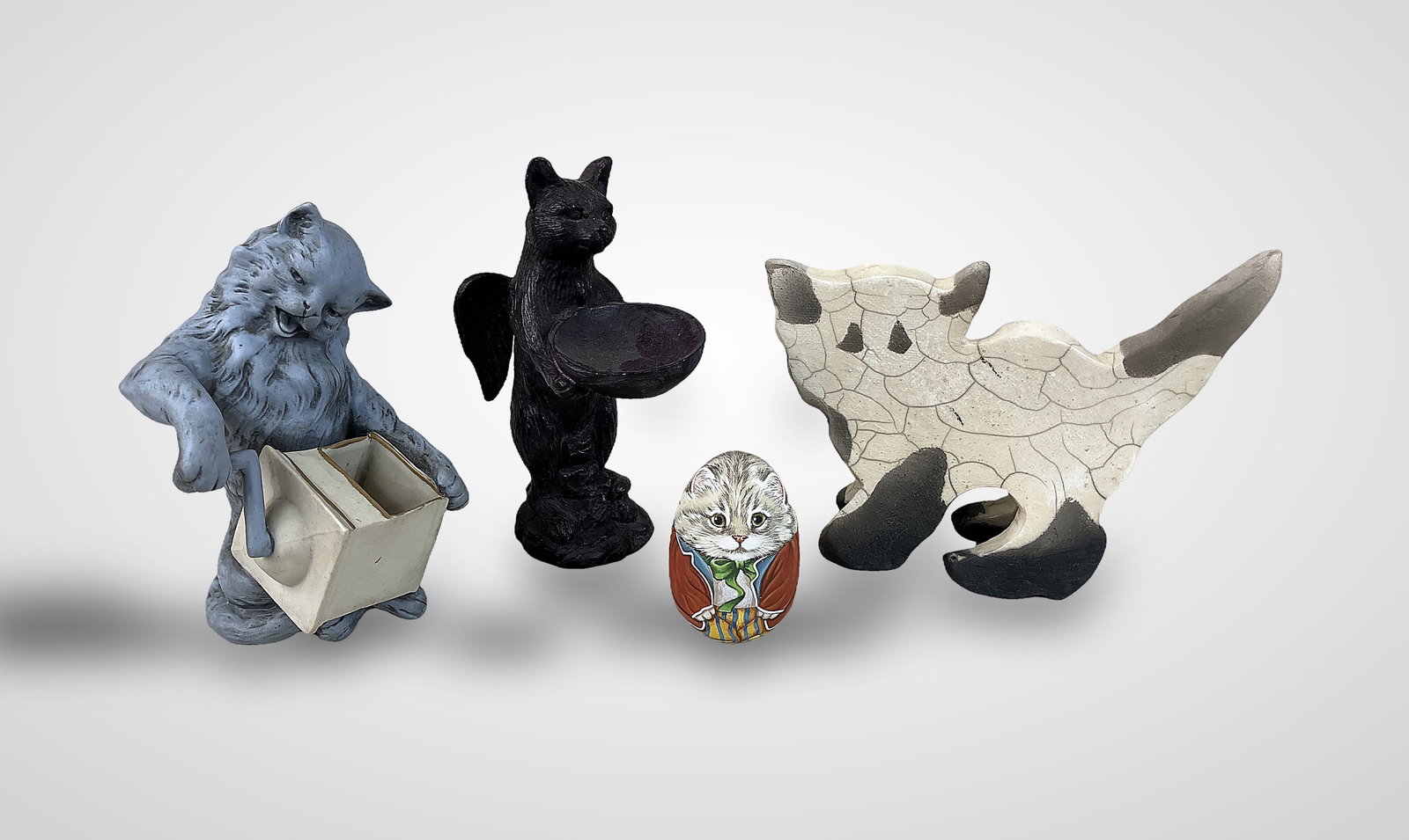 Four Cat Figurines: Eclectic group includes four distinct items: a blue ceramic piece depicting a cat turning a wind-up toy box; a raku-glazed ceramic figurine; a dark resin trinket dish featuring a standing cat angel, m