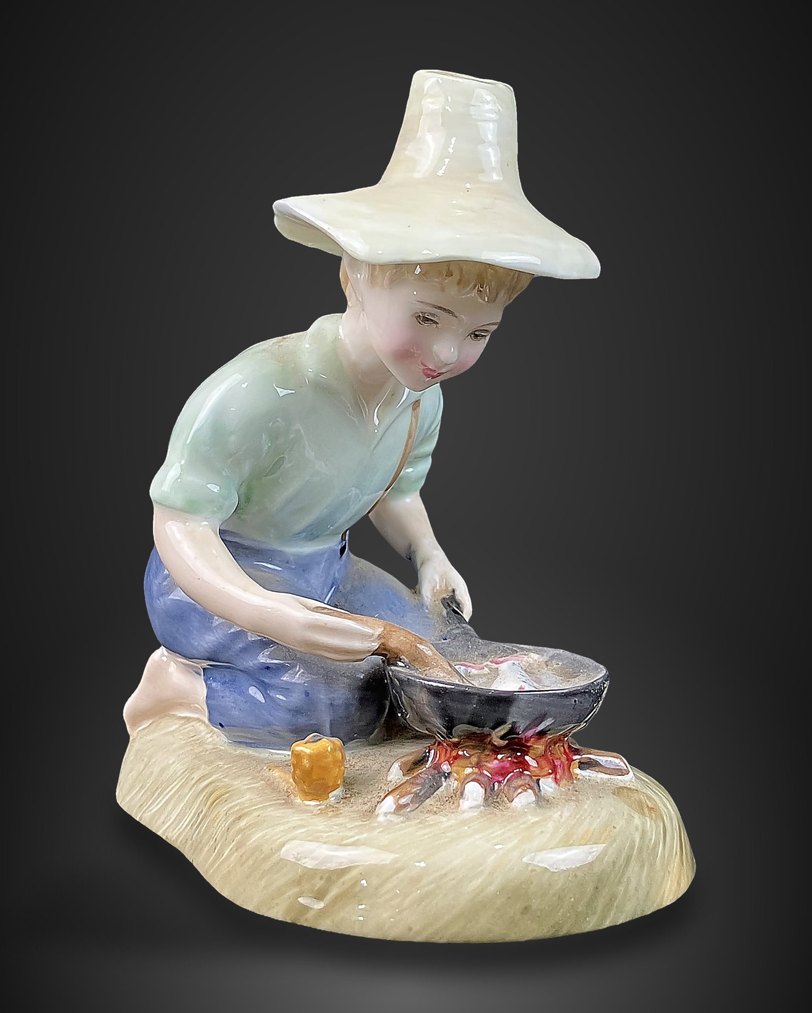Royal Doulton River Boy Figurine: Mid-century porcelain piece was designed by Peggy Davies and features a hand-painted scene of a boy in a straw hat cooking fish over a campfire. It measures approximately 4 in. H x 3 1/8 in. W x 2