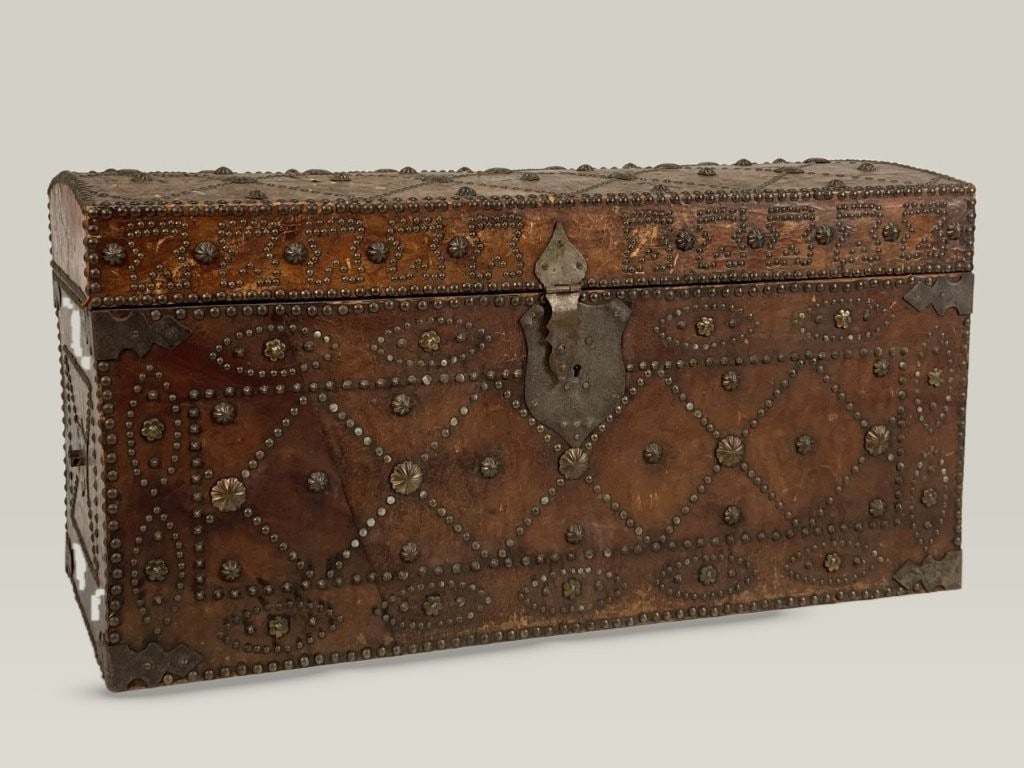 Spanish Revival Decorated Leather Trunk: Spanish colonial-style domed trunk features a rich, warm leather-wrapped form accented with nail heads and decorative metal elements that create geometric patterning across the exterior. Fitted with