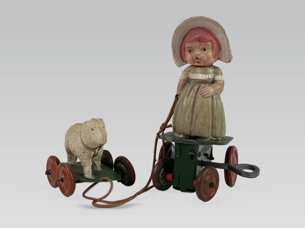 20th Century Mary and Lamb Wind-Up Toy: This 20th-century wind-up toy is crafted from celluloid and tin. It features a green wind-up base for the figure of Mary and a separate wheeled platform for the lamb, with the two connected by a