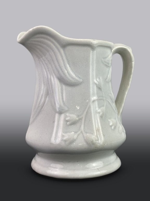 Ironstone Pitcher: 19th-century piece features a pristine white form accented with a floral relief design in the style of bellflowers. It measures approximately 8 in. H x 7 1/4 in. in diameter, including the handle.