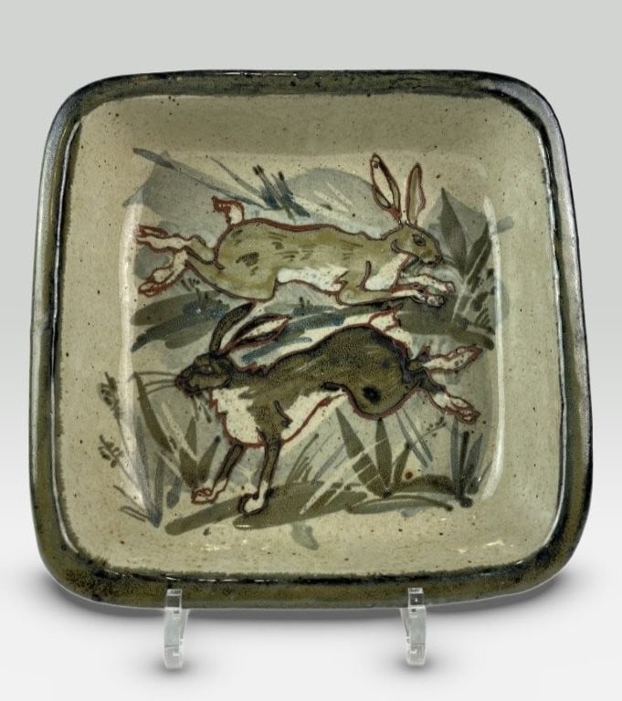 Maureen Minchin Pottery Rabbit Casserole Dish: This pottery casserole dish, created by Maureen Minchin, features a scene of two rabbits in a running pose amongst foliage. The design is rendered in rich green tones with red-colored outlines around