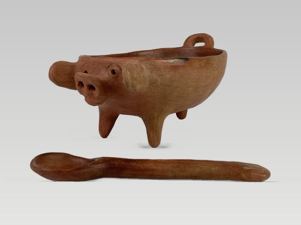 Mexican Folk Art Terracotta Pig Dish: This terracotta dish is sculpted in the form of a pig, featuring a dramatically elongated snout, protruding ears, and three small legs. It includes a wooden spoon and measures approximately 4 1/2 in. 
