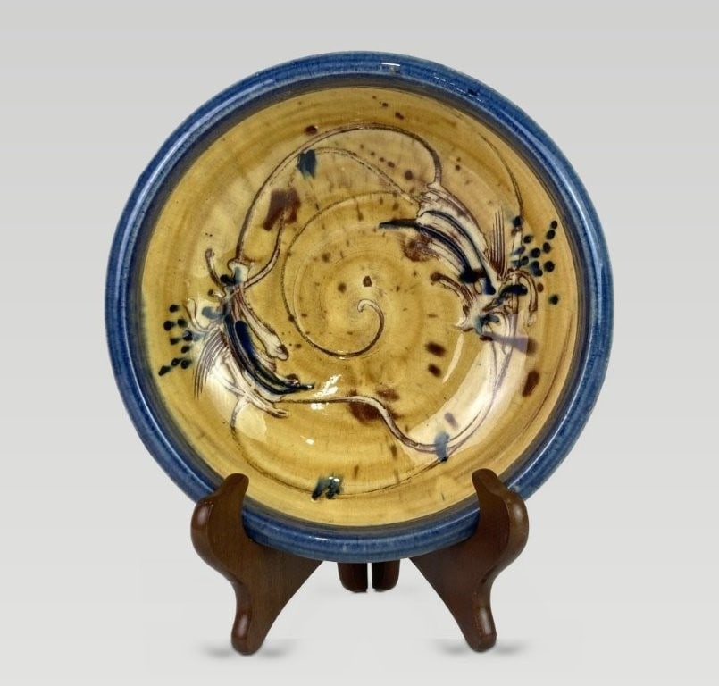 Studio Art Pottery Bowl: This bowl features a vibrant yellow glaze with a contrasting blue rim. The interior displays abstract, organic flowing forms in shades of brown, blue, and cream, which continue along the exterior side