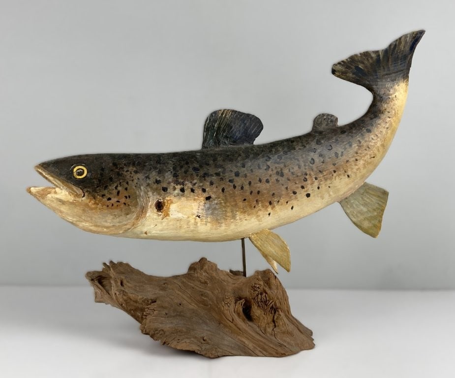 Folk Art Brown Trout Wood Sculpture: This handcrafted wood sculpture depicts a dynamic brown trout with its tail swooping into the air and a naturalistically spotted body, securely mounted to a weathered driftwood base. Displays a worn