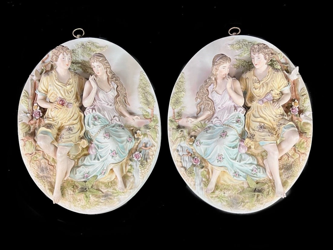 Oval Bisque Porcelain Wall Plaques Courting Couple: Pair of Oval Bisque Porcelain Wall Plaques with courting couples, surrounded by flowers and birds, all dimensional. Makers mark on reverse. Metal hanger. Measurements: Approx. 12.75 x 10.25 x 4 inches