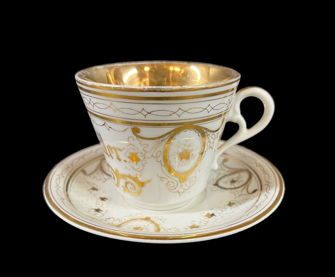 KPM Porcelain Cup and Saucer c 1880: KPM Porcelain Teacup and Saucer c 1880, hand painted gilt designs. Saucer reads, 'Bum Andenken den 14 Mai 1882 von A. F.', the cup 'Julius Muller.' Makers mark on underside, underglaze blue. Measureme