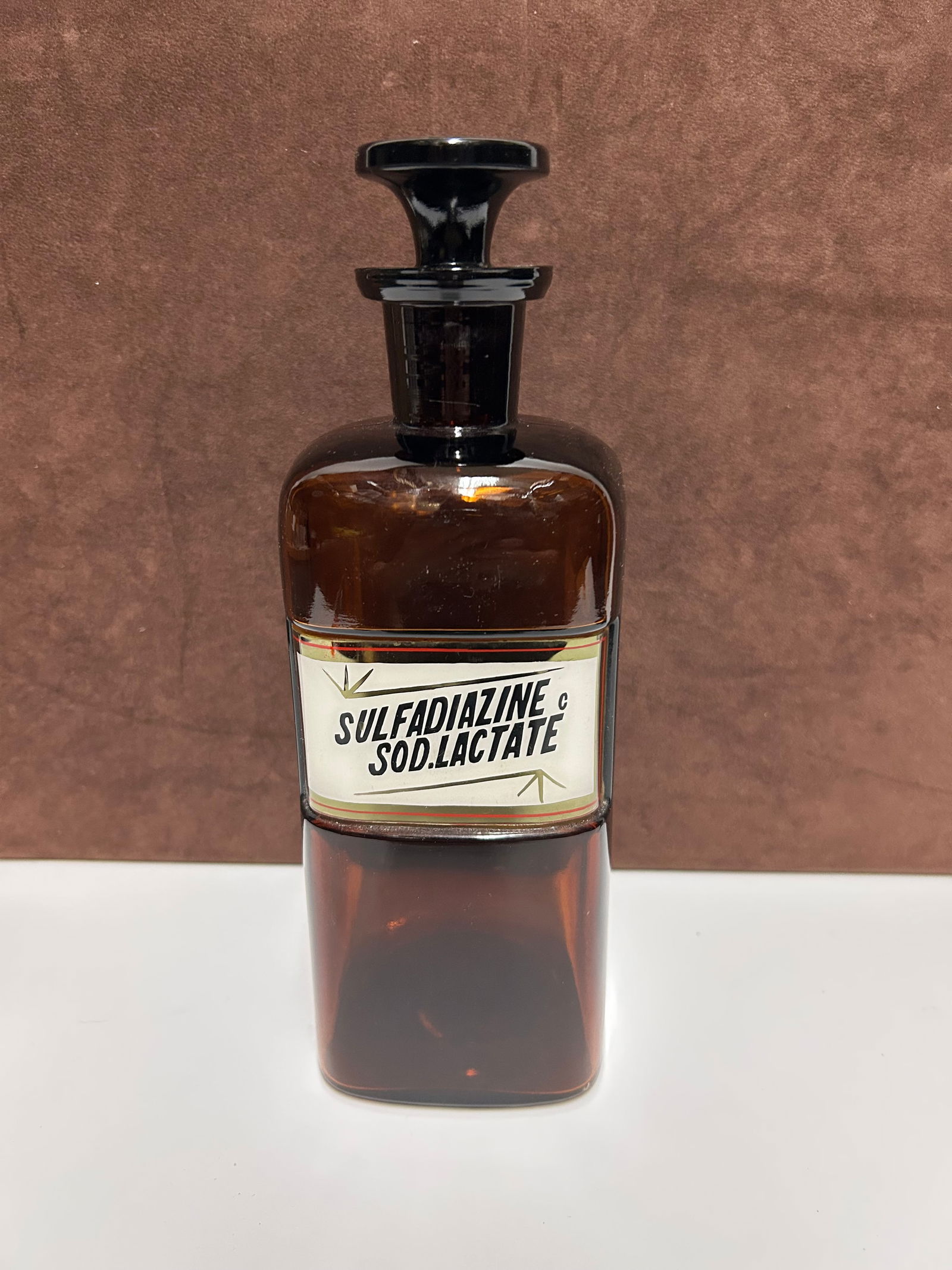 Amber Glass Apothecary Bottle Labeled “SULFADIAZINE SOD.LACTATE”: This vintage amber glass apothecary bottle dates to the early 20th century, during a period when pharmacies transitioned from traditional herbal preparations to scientifically formulated medicines. Cr