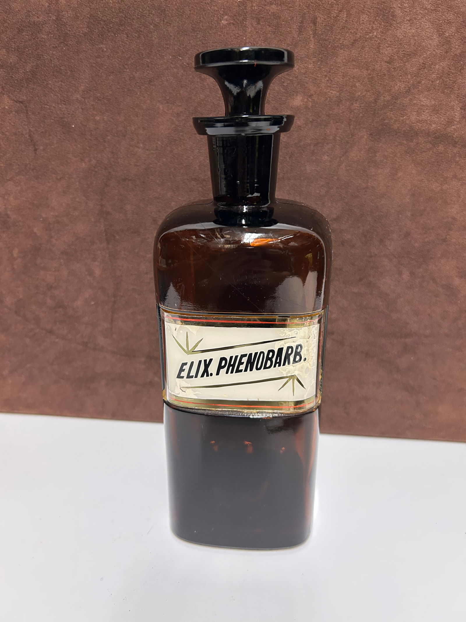 Amber Glass Apothecary Bottle Labeled “ELIX. PHENOBARB.”: This antique amber glass apothecary bottle measures 10 inches tall by 3 inches wide and dates to the late 19th to early 20th century. It once held Elixir Phenobarbital, an early sedative preparation u