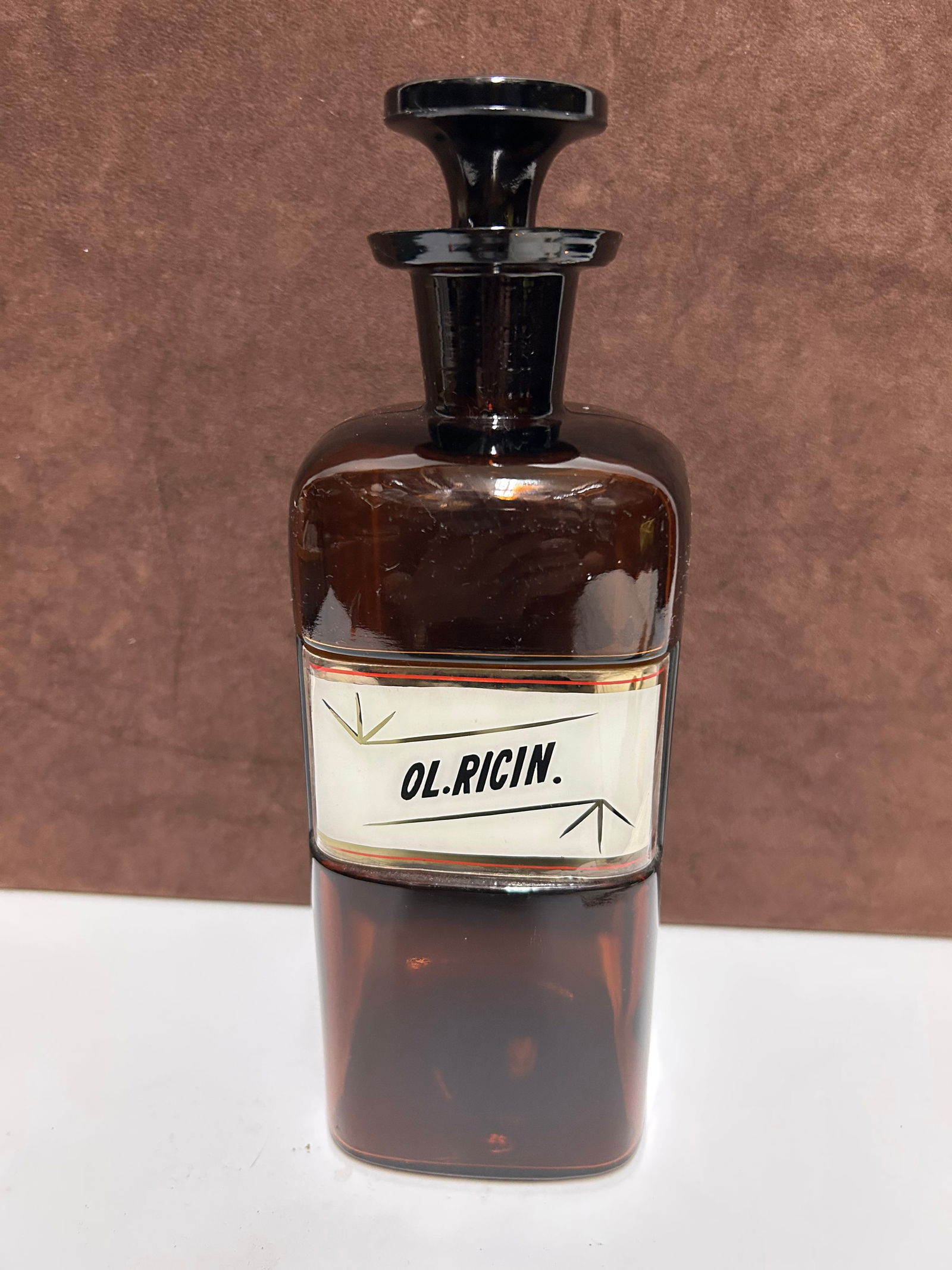 Amber Glass Apothecary Bottle Labeled “OL. RICIN.”: This antique amber glass apothecary bottle measures 10 inches tall by 3 inches wide and dates to the mid-to-late 19th century, circa 1850–1890. Designed for pharmacy use, it features deep amber glas
