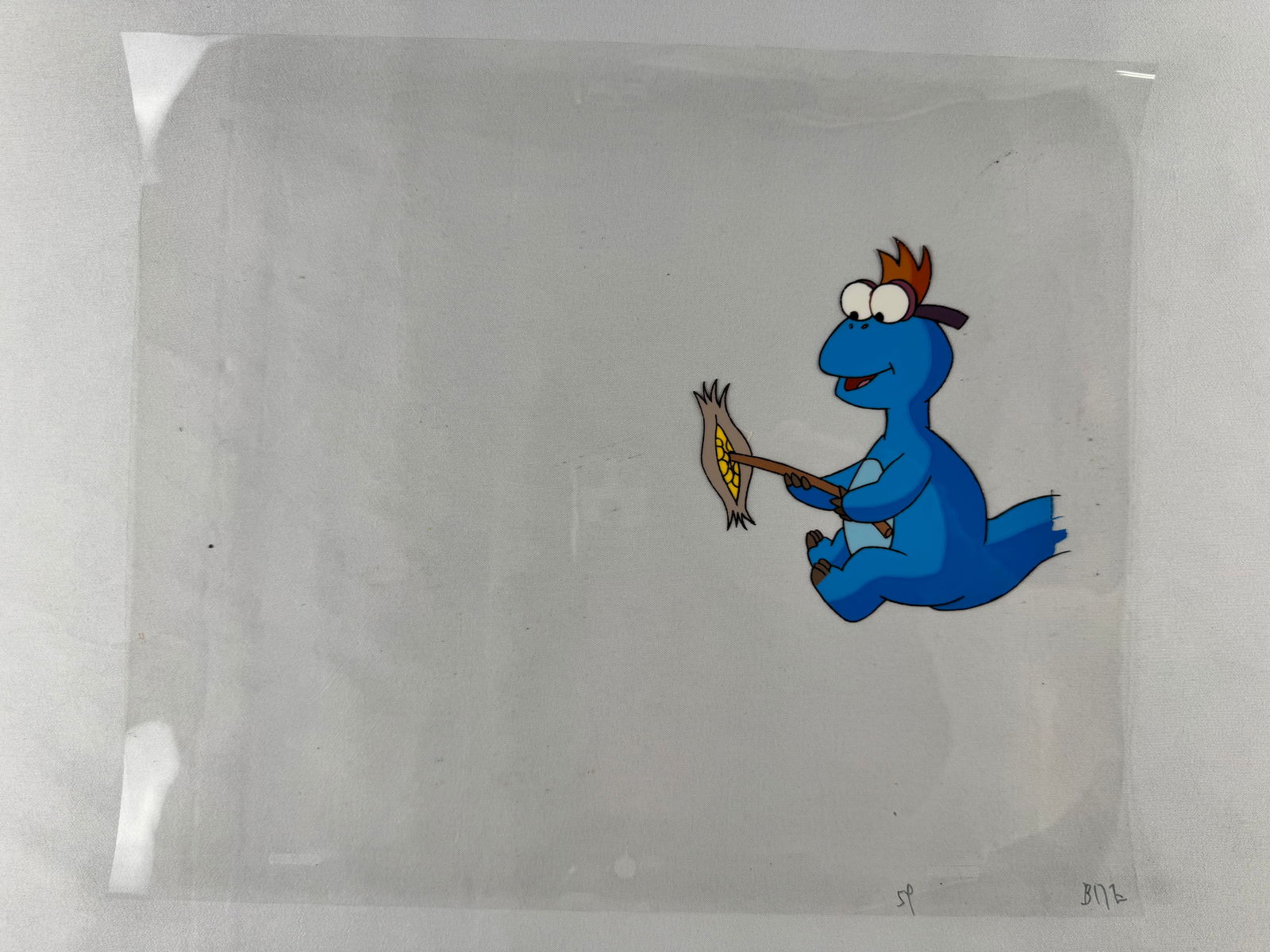 Animation Cel, Truman from Dino Babies Holding Corn Cob: Animation cel featuring a blue cartoon creature seated and holding a yellow corn cob. The cel is photographed on a clear sheet over a neutral background.