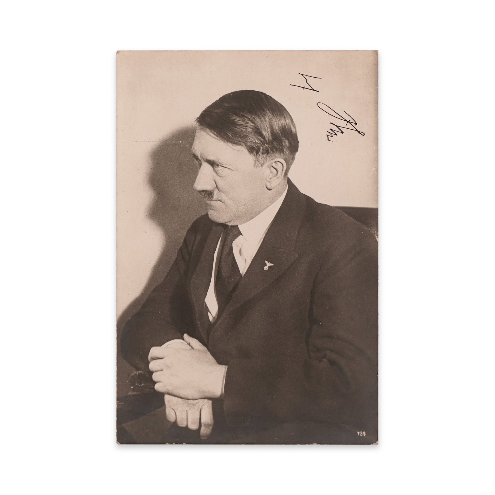 ADOLF HITLER SIGNED POSTCARD: This signed photographic postcard captures Adolf Hitler in a seated three-quarter profile, with hands clasped and gaze directed forward in a contemplative posture. The sharp studio lighting casts a di