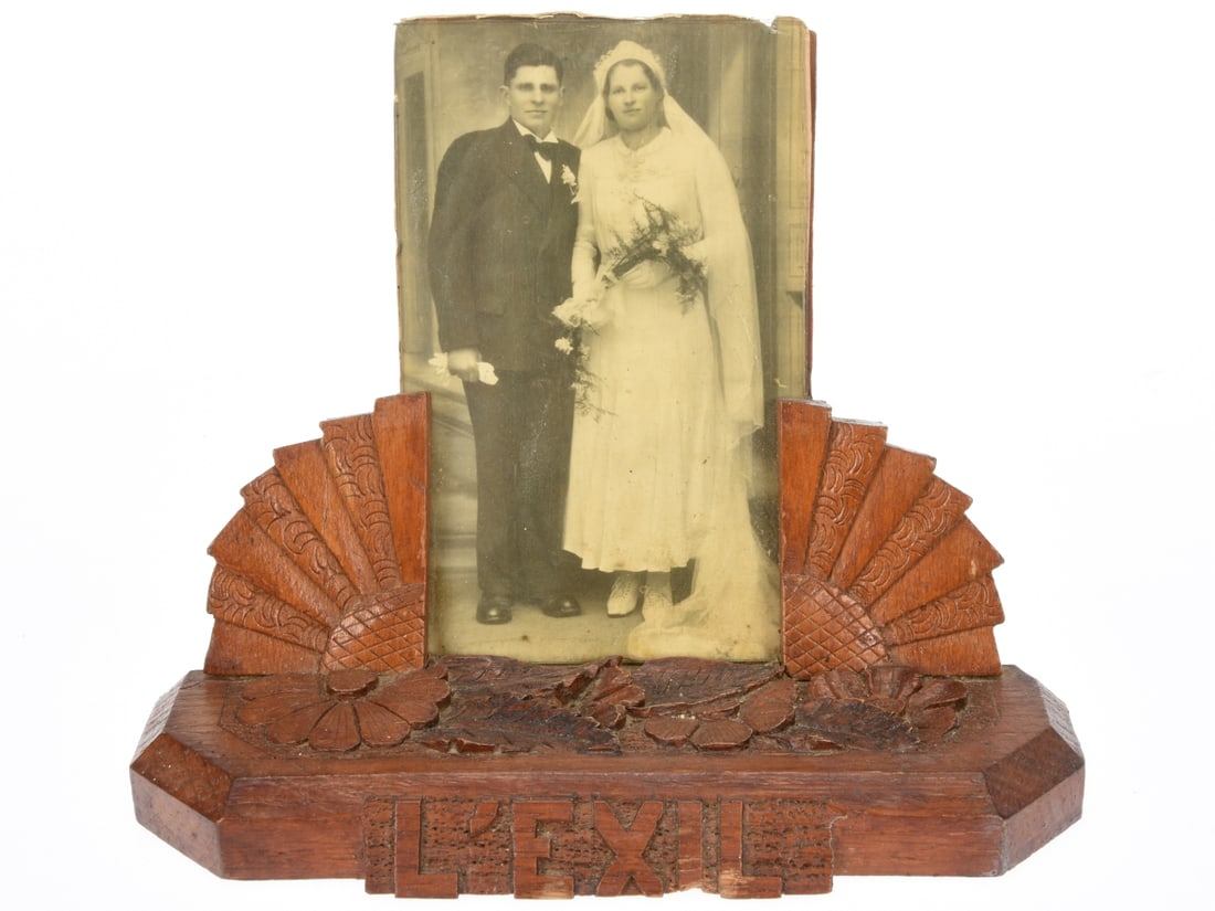 French Interned Refugee Picture Frame: 8" long wooden base is skillfully chip carved with flowers and leaves on the top surface. L'EXIL is carved into the front edge. Two carved uprights resembling quarters of a sunflower support the weddi