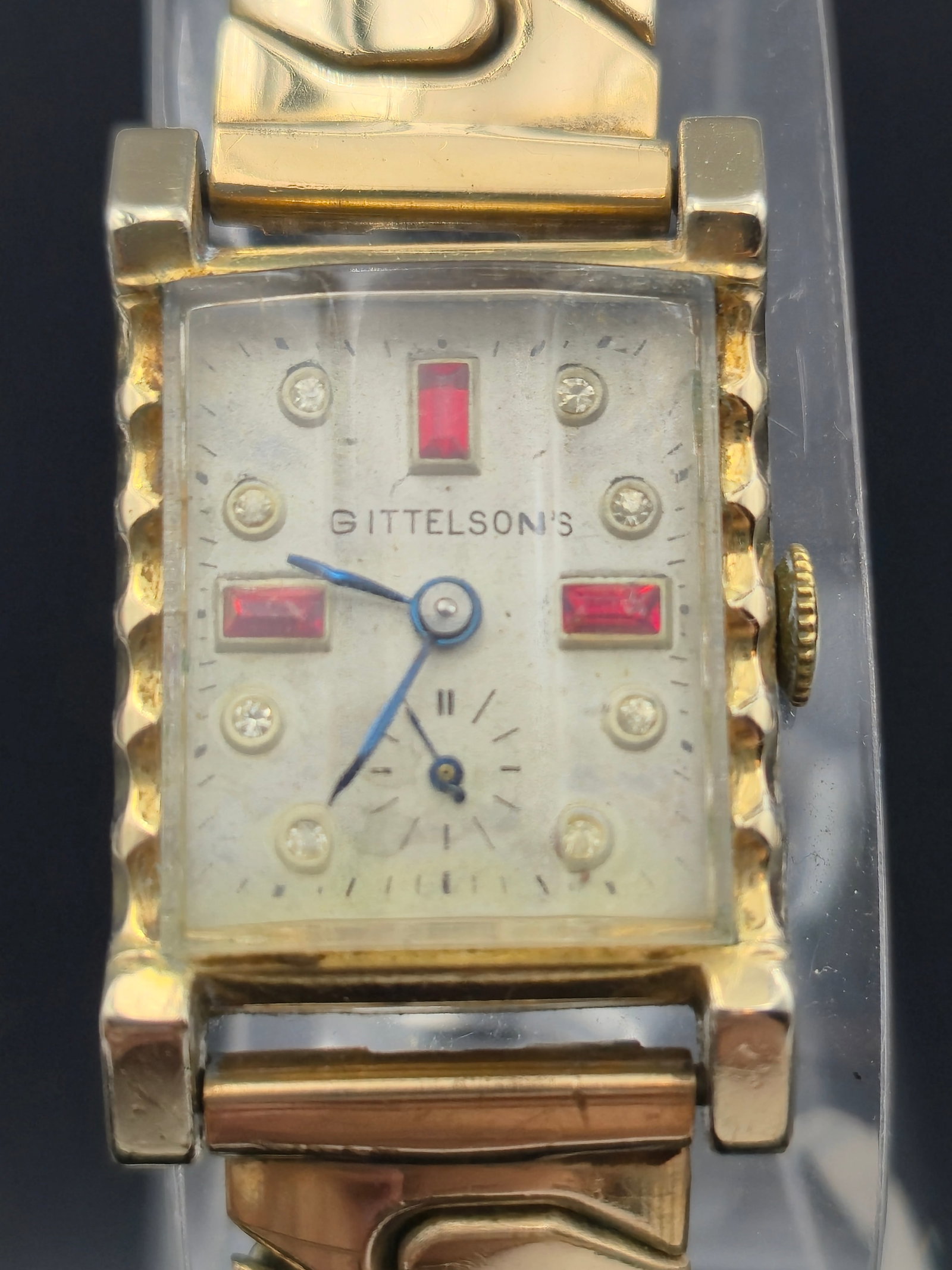 1940s A.Hirsch 7J 10k RGP art deco mens tank watch: This elegant Gittelson’s vintage wristwatch features a striking rectangular gold-tone case with Art Deco styling and decorative band links. The dial is accented with red and clear stone markers, blu