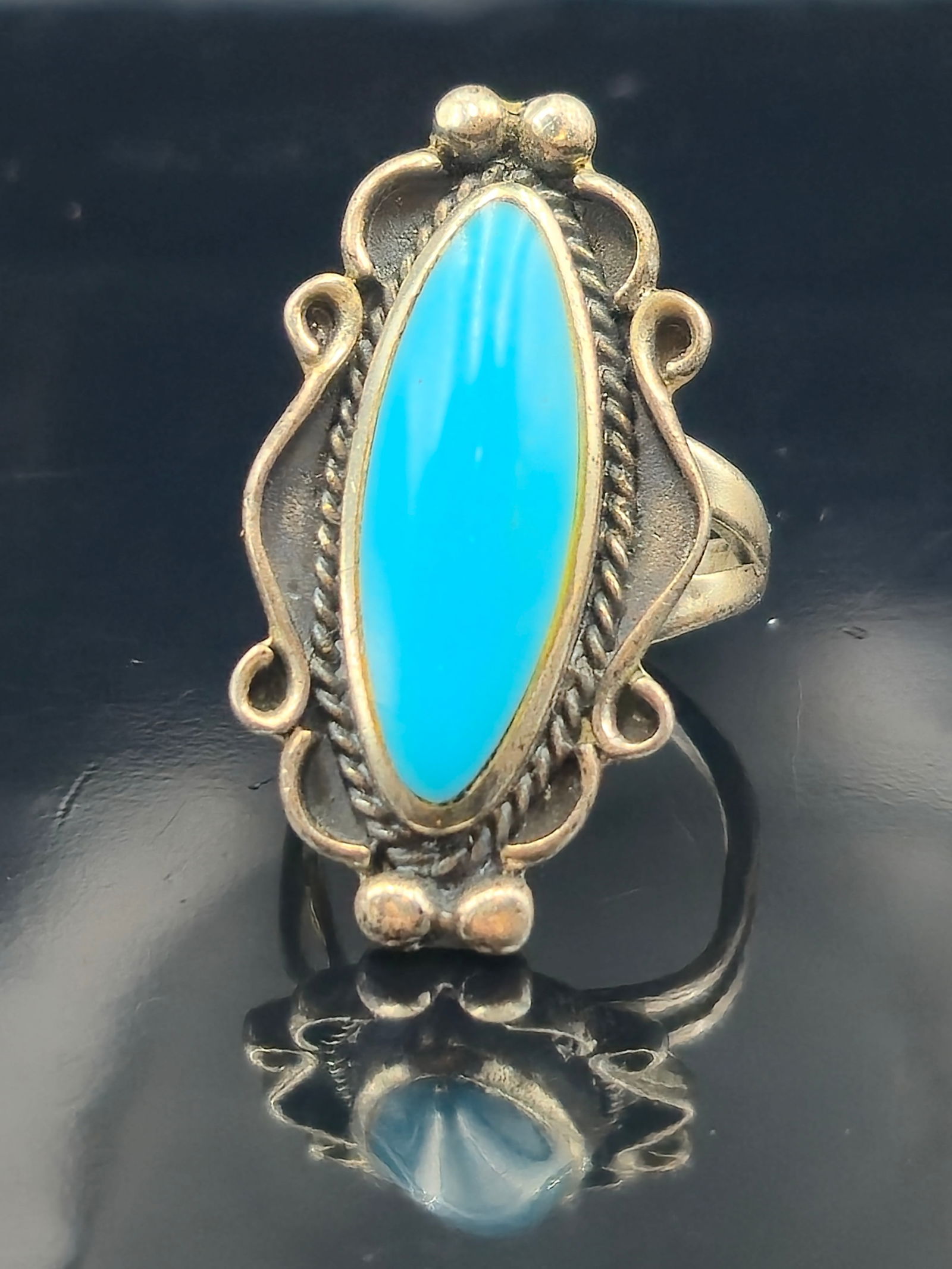 Navajo Sterling Long Turquoise Ring Sz 5: Vintage Navajo (Diné) sterling silver ring featuring a long oval turquoise cabochon in a twisted rope bezel, framed by applied silver scrolls and raindrop beads. Marked STERLING on reverse. Classic S