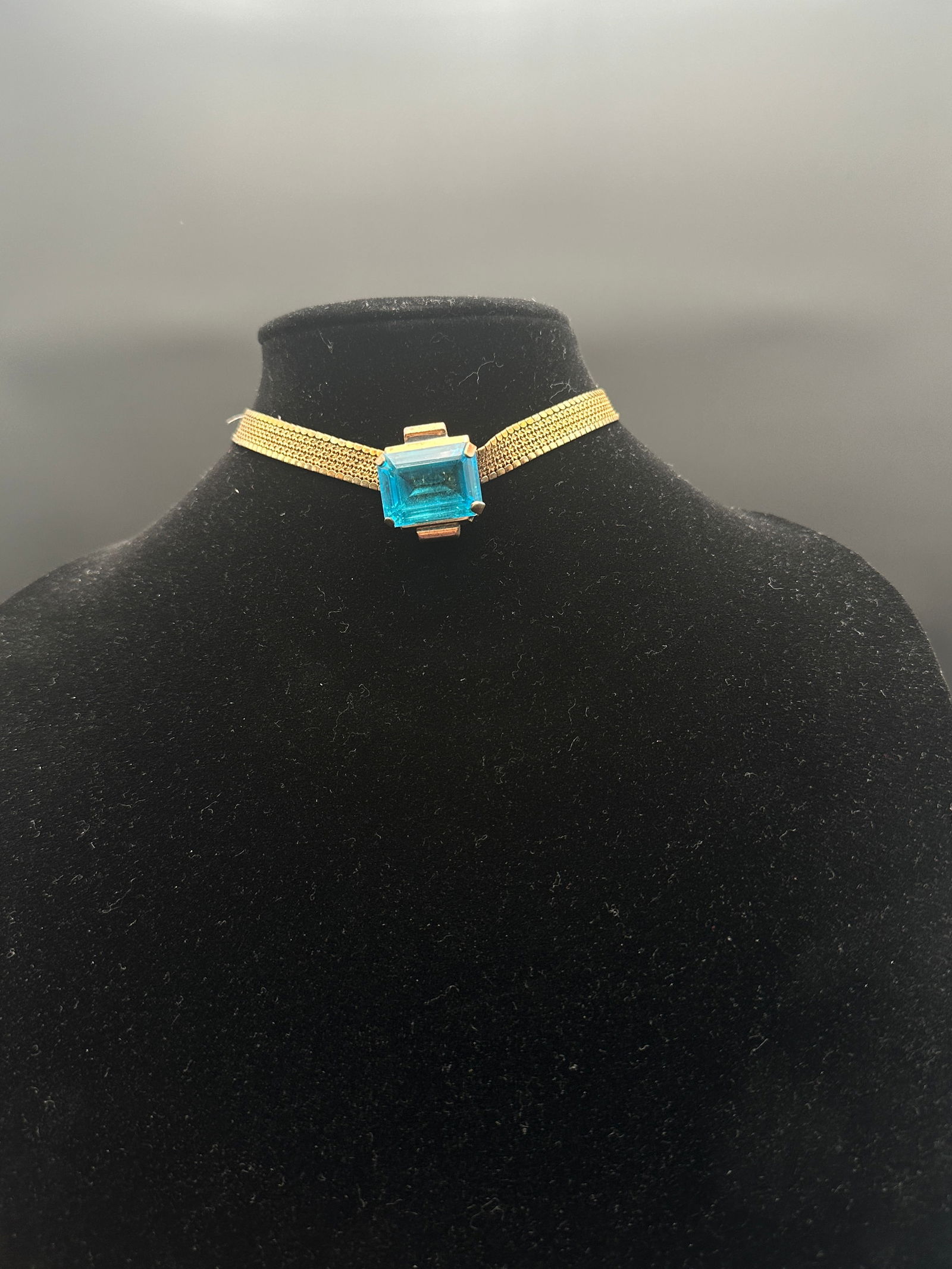 Vintage Gold Tone Mesh Choker Necklace with Large Blue Emerald-Cut Glass Stone: A striking vintage gold tone choker necklace featuring a wide mesh chain with a bold, emerald-cut blue glass centerpiece in a four-prong setting. The vibrant aqua-blue stone catches the light beautifu
