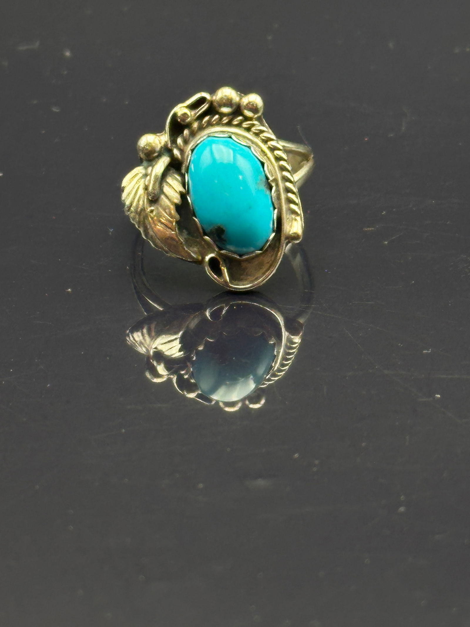 Vintage Sterling Silver Turquoise Ring with Leaf & Bead Accents: A vintage sterling silver ring featuring a vivid turquoise cabochon set in a bezel, framed with rope detailing and accented by a silver leaf and bead motifs. Marked Sterling on the reverse. Classic So