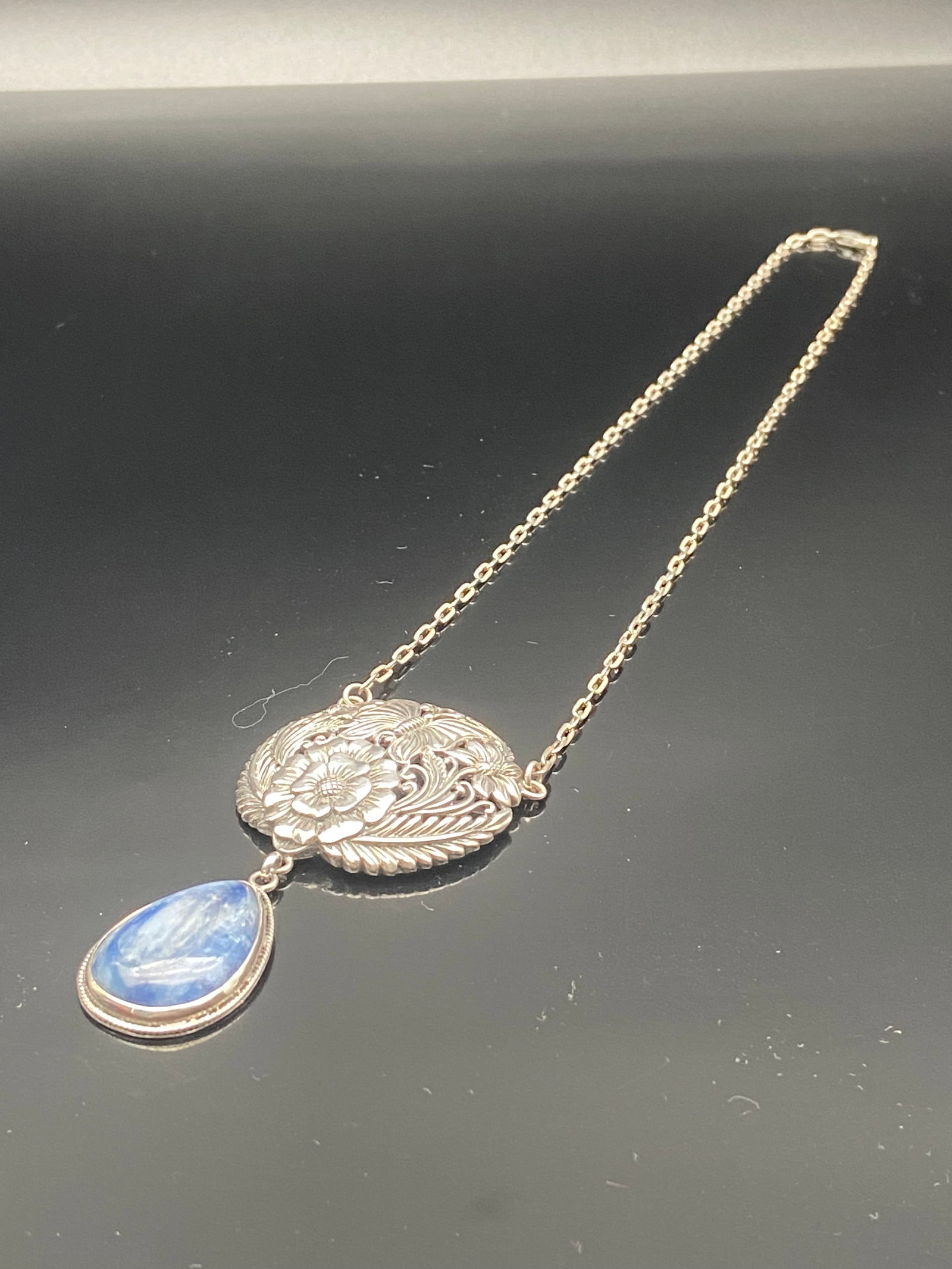 Sterling Silver Floral Necklace w/ Blue Stone Drop: Elegant sterling silver necklace featuring a detailed floral medallion with engraved leaf and blossom motifs. Suspended from the medallion is a pear-shaped polished blue cabochon stone, bezel-set in s