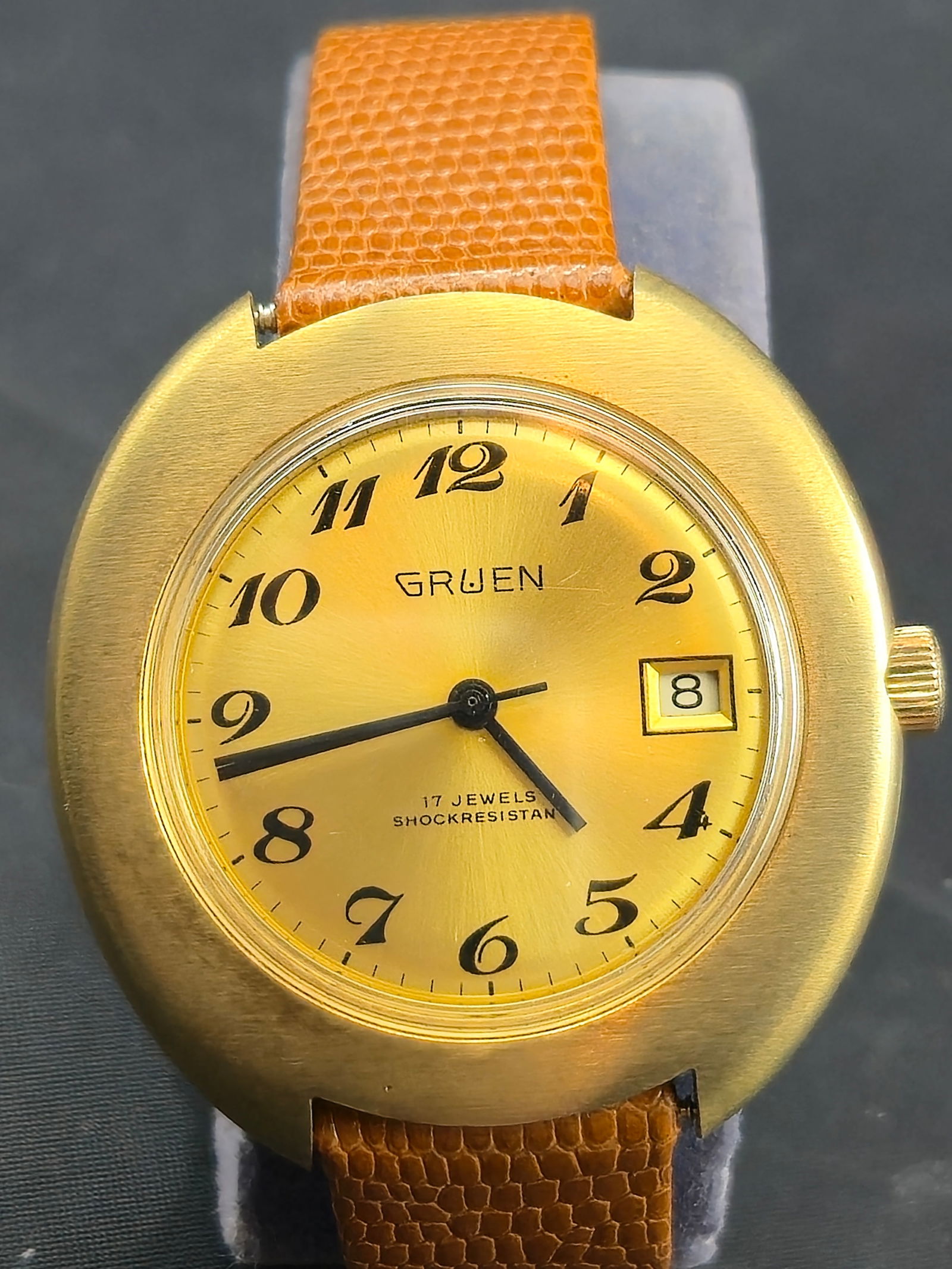 1970s Gruen Mechanical Wristwatch Gold-Tone Cushion Case – 40mm: This vintage Gruen wristwatch from the 1970s features a bold retro cushion-style case in gold tone, paired with a matching sunburst gold dial. The dial is accented by applied black Arabic numerals, sl
