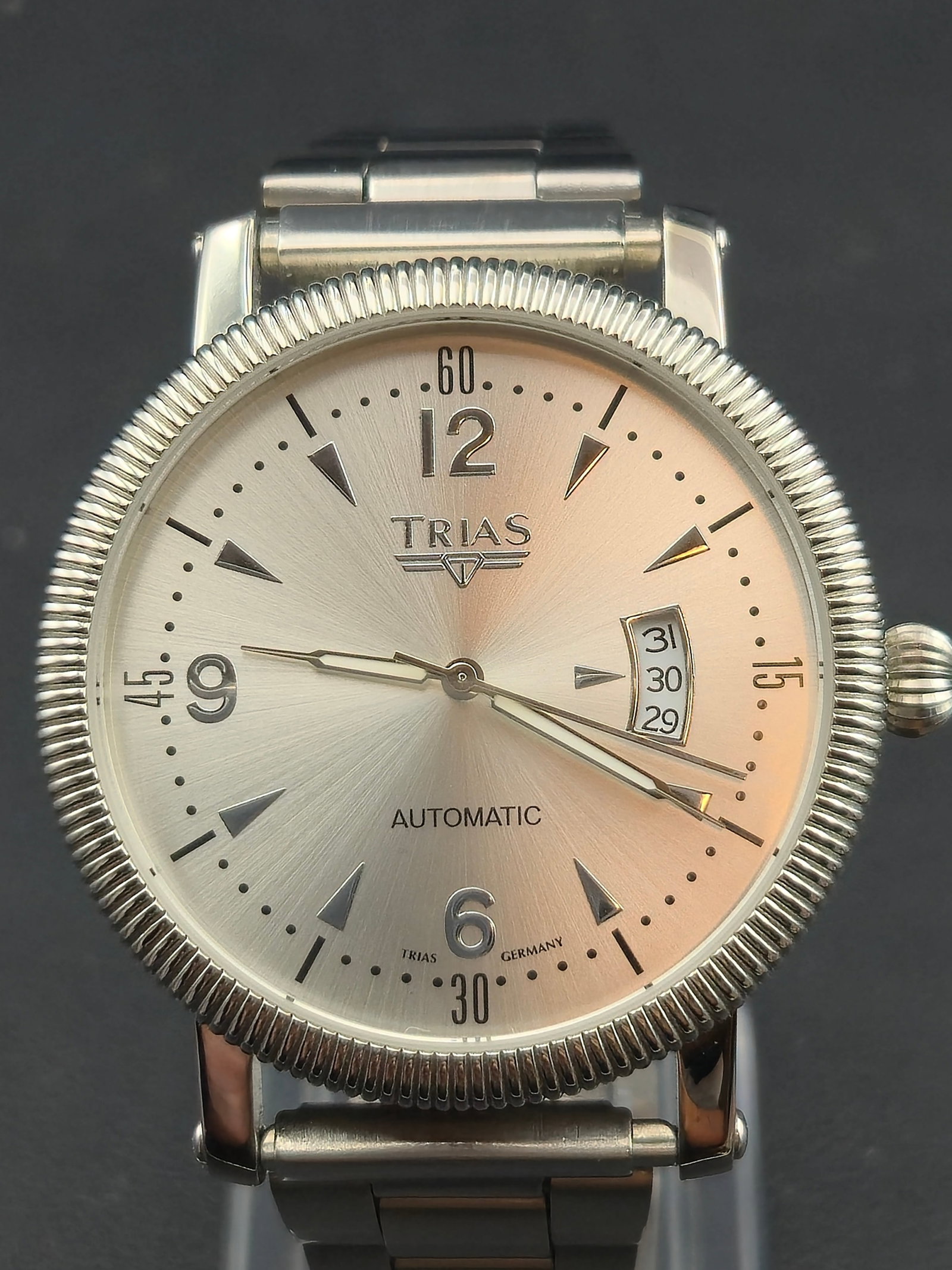 Trias Ref. 01064 Germany Automatic Silver Dial Wristwatch (41mm, Running): Elegant Trias Germany automatic wristwatch featuring a finely brushed silver sunburst dial with applied Arabic numerals, luminous hands, and a unique curved date display between 2 and 4 o’clock. The