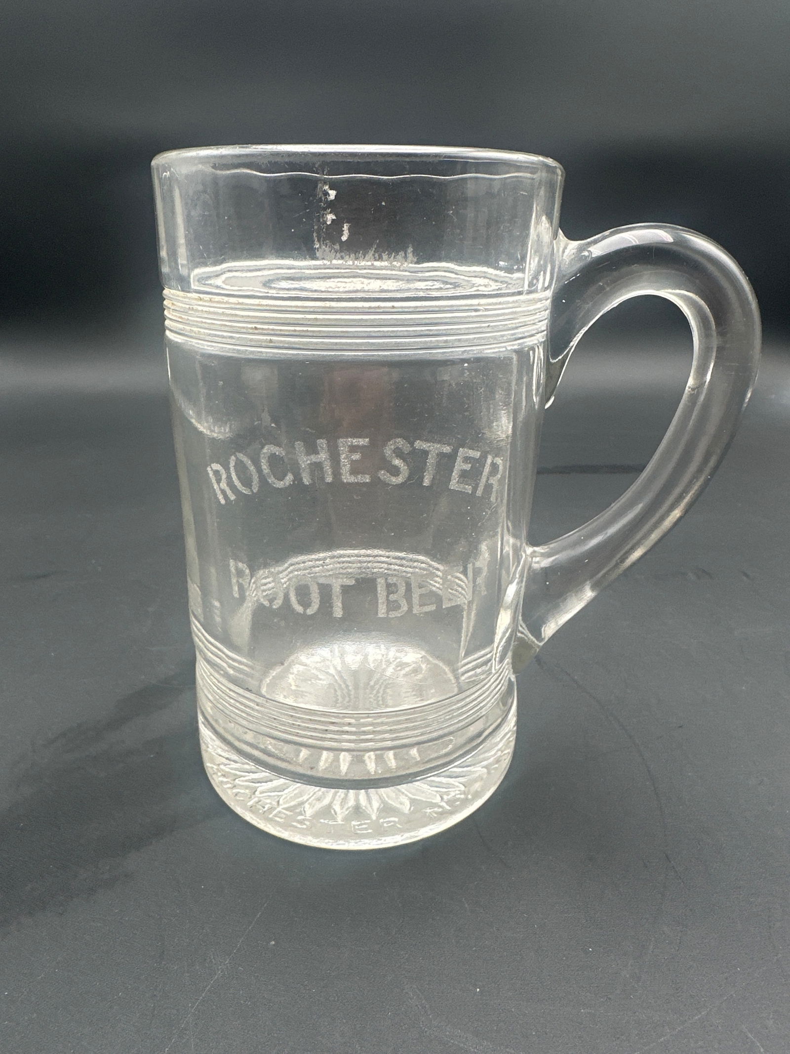 Vintage Rochester Root Beer Advertising Glass Mug: Heavy clear glass root beer mug featuring etched “Rochester Root Beer” logo, ribbed detailing, and sunburst base. Classic soda fountain advertising collectible from mid-20th century America. E