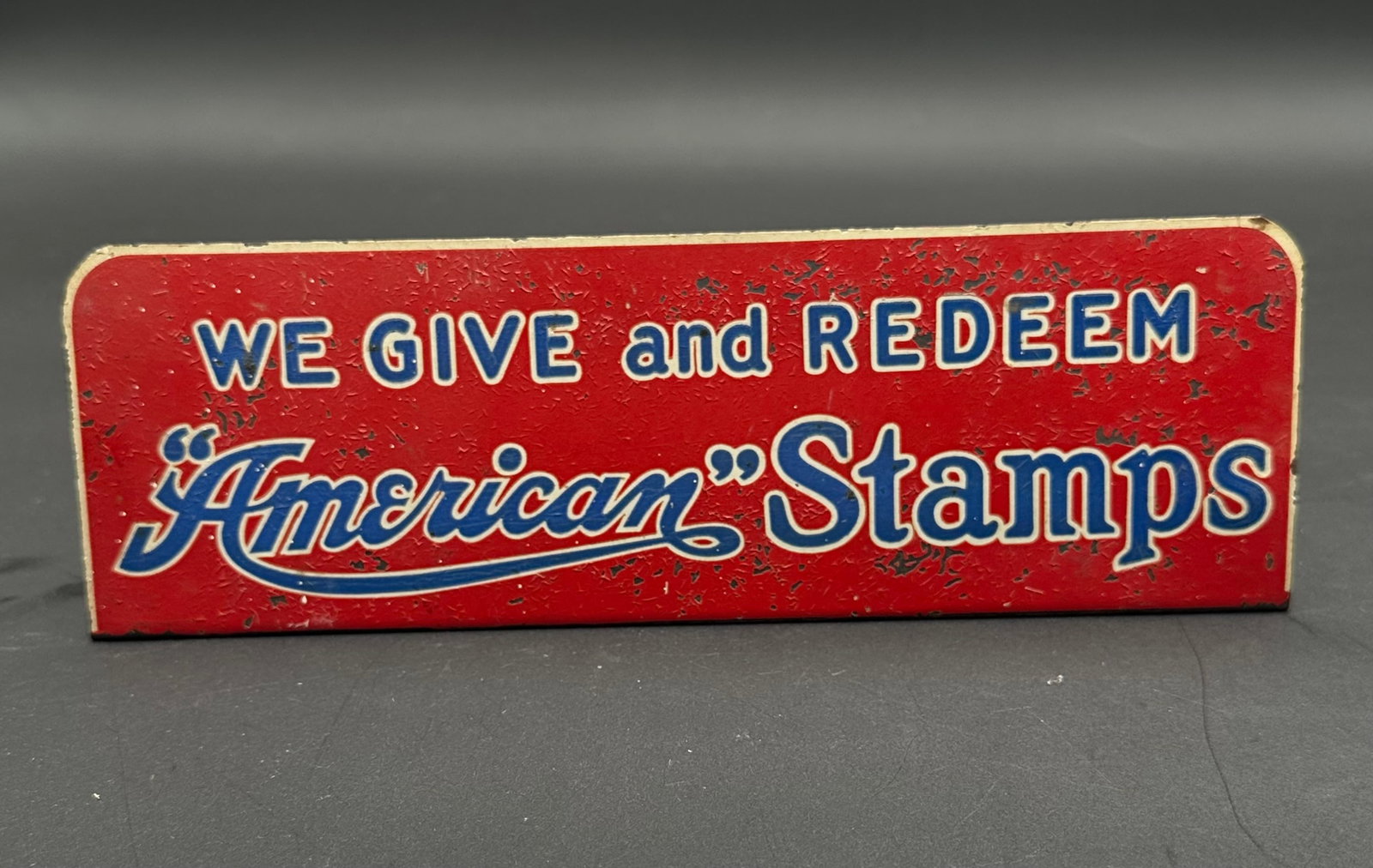Vintage “American Stamps” Advertising Counter Sign: Original mid-century tin advertising sign reading “We Give and Redeem American Stamps.” Bright red background with bold blue and white lettering, made for in-store display by The American Stam