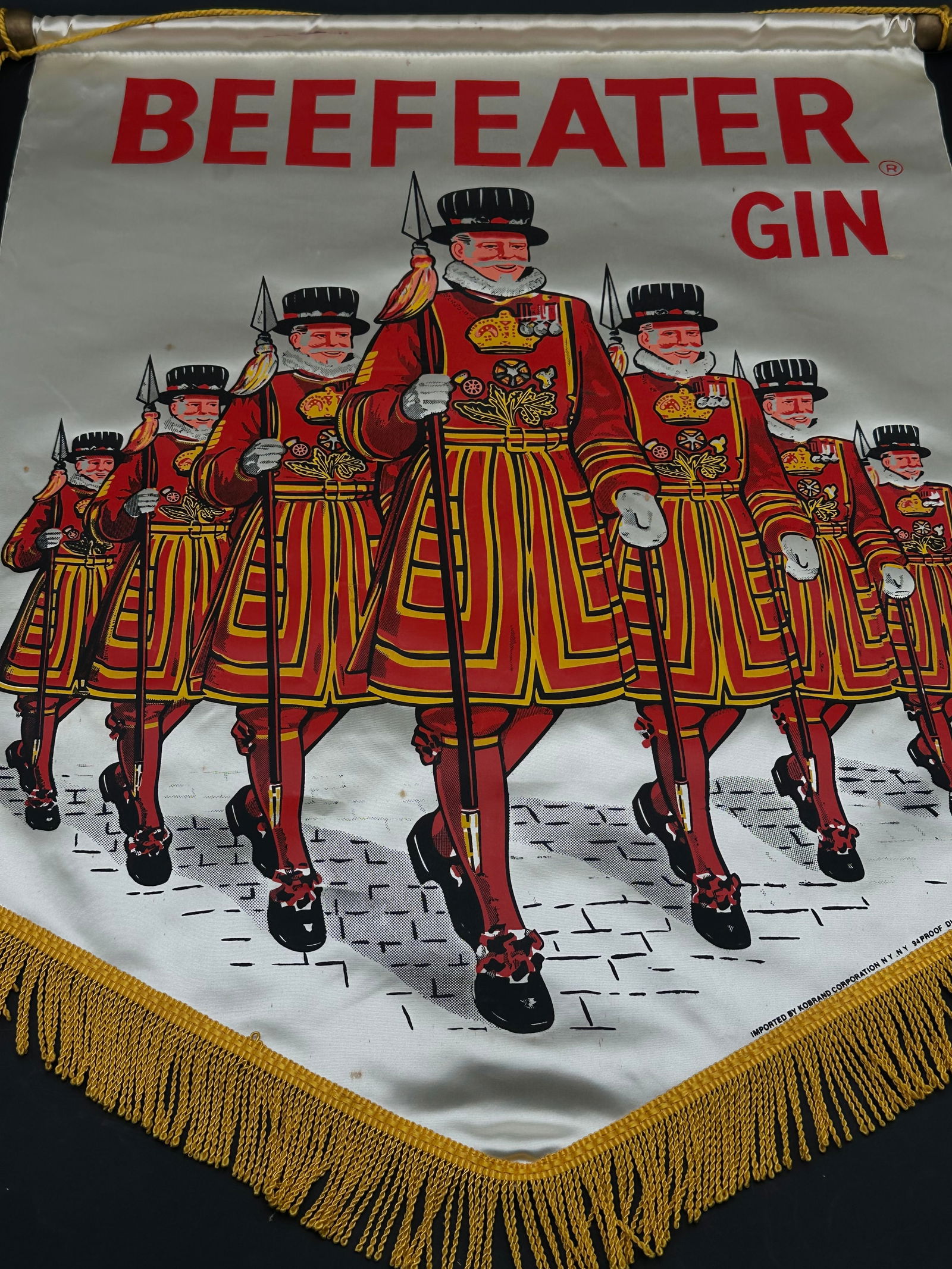 Vintage Beefeater Gin Advertising Banner with Beefeaters Parade: Colorful mid-century Beefeater Gin fabric banner featuring marching Yeomen Warders in bright red and gold uniforms. Made of satin-like material with gold fringe trim and wooden dowel. Displays vibrant