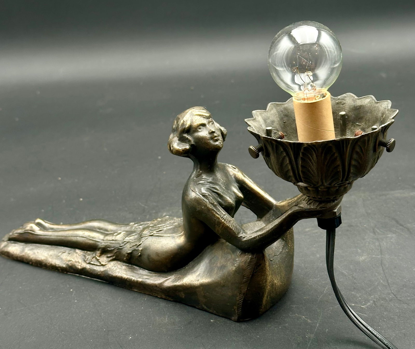 Art Deco Nude Woman Lamp with Floral Shade Holder: Elegant vintage Art Deco figural lamp featuring a reclining nude woman in cast metal with a bronze finish, holding a flower-form light socket. Ornate leaf and shell detailing around the shade holder.