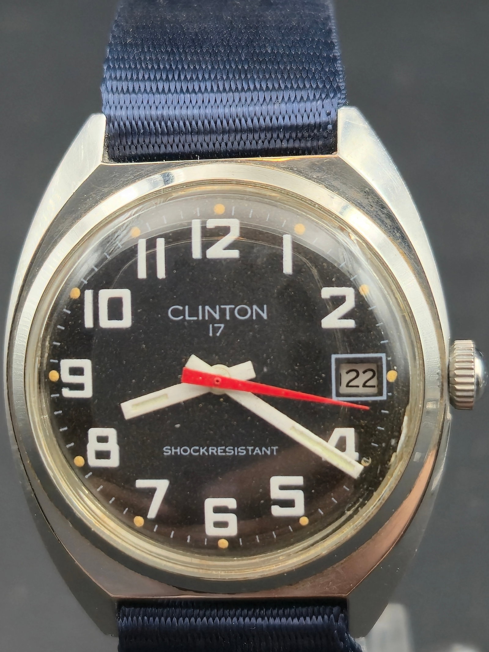 Clinton 17J Manual Wind 34 mm Military Style Date Watch – Runs: Vintage Clinton 17-jewel manual wind wristwatch with a classic black military-style dial featuring bold white Arabic numerals, red sweep seconds hand, and a framed date window at 3 o’clock. The stai