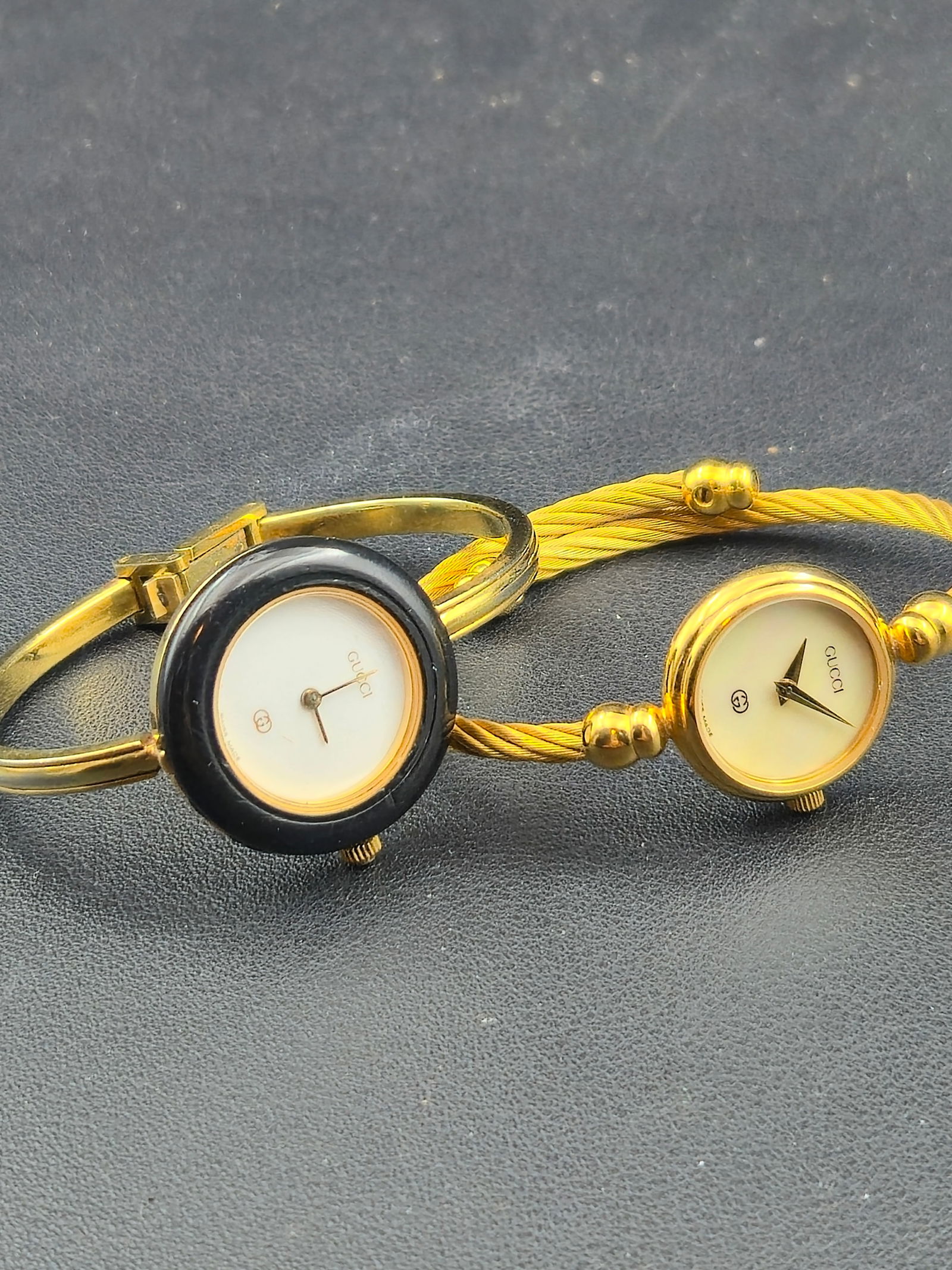Lot of 2 Gucci Ladies Swiss Watches – MOP Dial Running + 1100-L Black Bezel Untested – Swiss: Lot of two Gucci ladies Swiss quartz watches. The first features a mother-of-pearl dial with gold hands and a twisted cable bangle bracelet and is running. The second is a Gucci 1100-L black-bezel ban