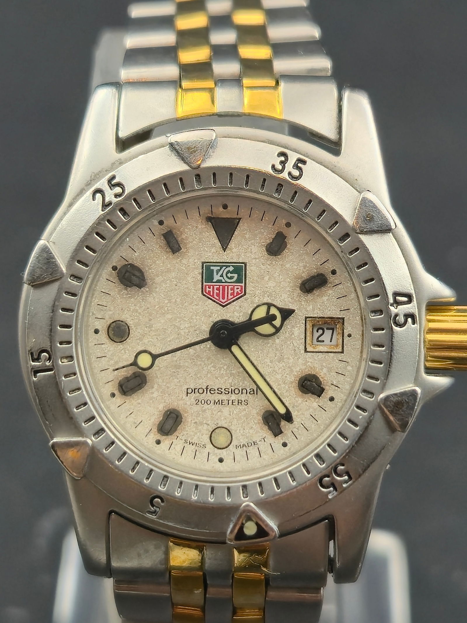 TAG Heuer Professional 200M Two-Tone 28mm Ladies Diver – 1990s: This two-tone TAG Heuer Professional 200M features the classic 1990s diver styling with gold-accented crown, bezel riders, and integrated jubilee bracelet. The champagne textured dial has developed a