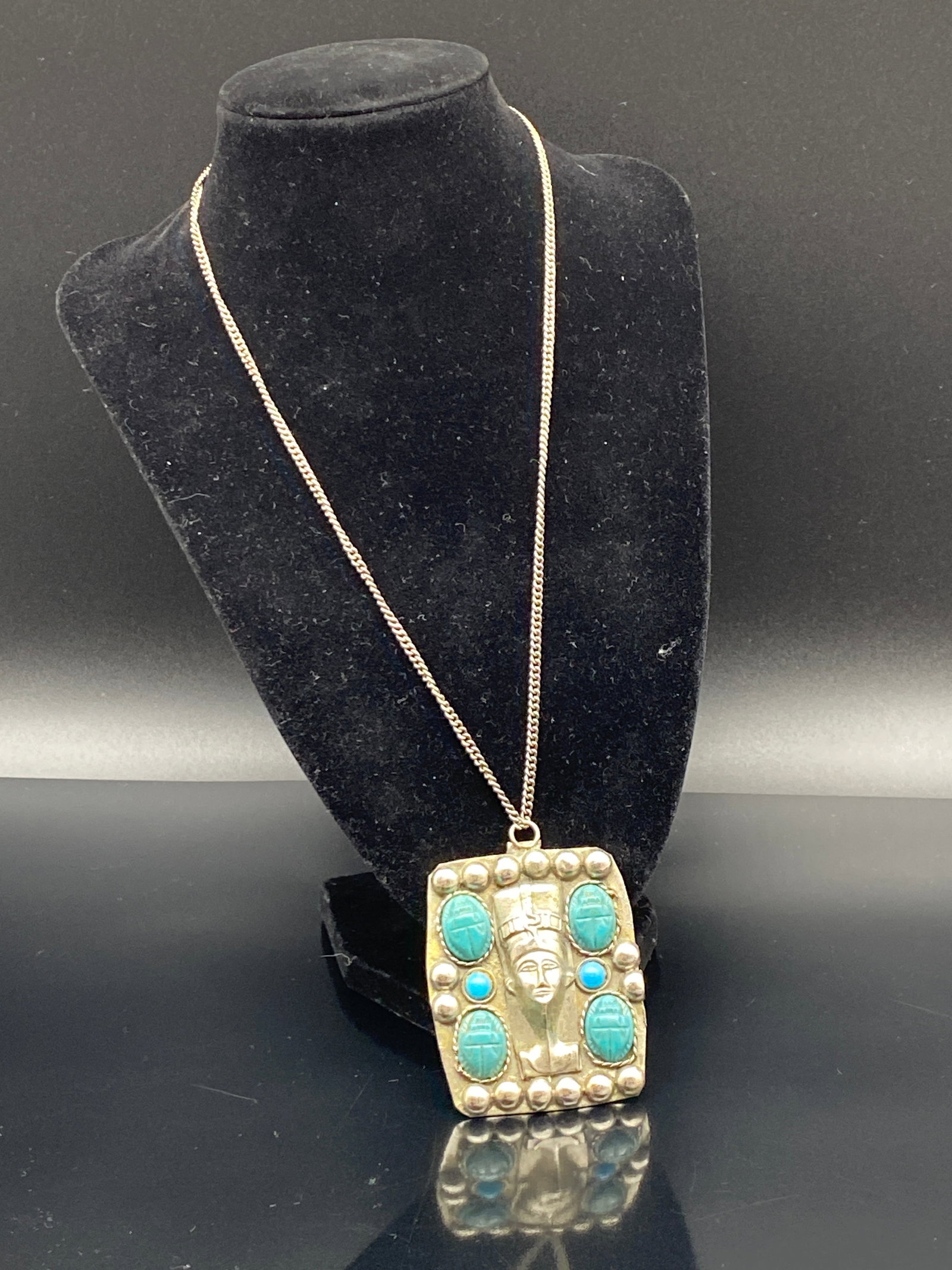 Egyptian Revival Turquoise-Style Pendant Necklace: A bold Egyptian Revival–style necklace featuring a large rectangular pendant with a raised pharaoh or queen’s bust at the center. The pendant is framed by rounded metal bead accents and set with f