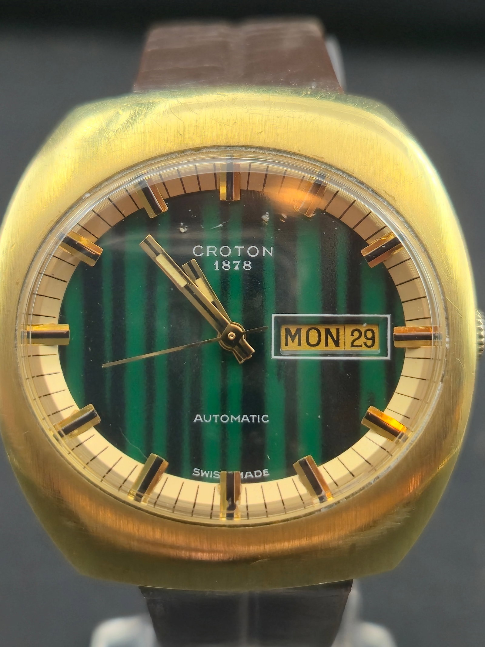 1970s Croton 1878 Automatic Day-Date Cushion Case Green Dial 37.2mm Swiss: Vintage 1970s Croton 1878 automatic wristwatch featuring a gold-tone cushion-shaped case and distinctive green vertical striped dial. Applied gold-tone hour markers with framed day-date window at 3 o�