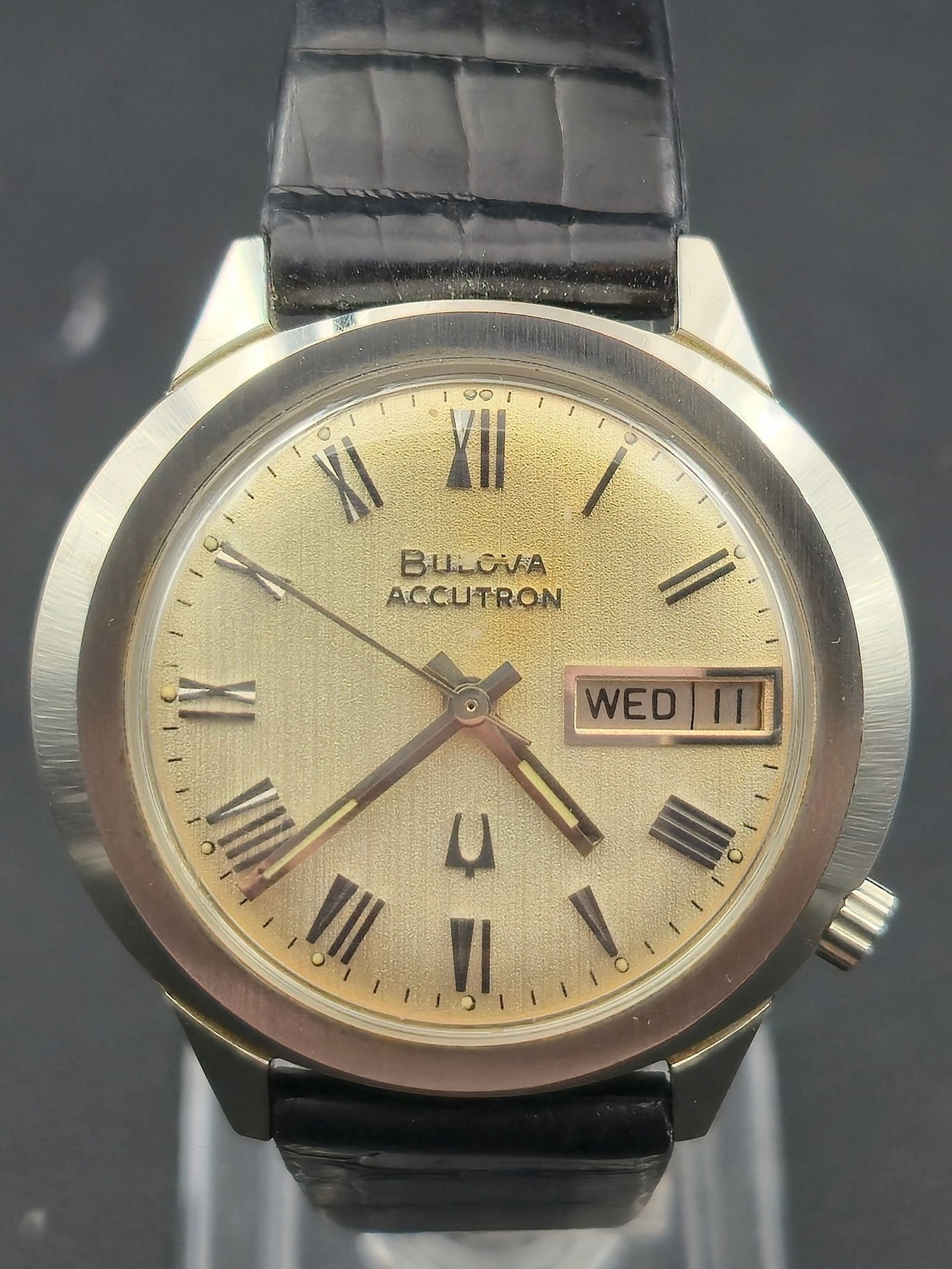 1969 Bulova Accutron 2182 Stainless Steel Case Champagne Dial Roman Numerals 34.0mm: Bulova Accutron wristwatch featuring a champagne-tone textured dial with applied Roman numeral hour markers and tuning fork logo. Day-date window at 3 o’clock displaying WED 11. Stainless steel case