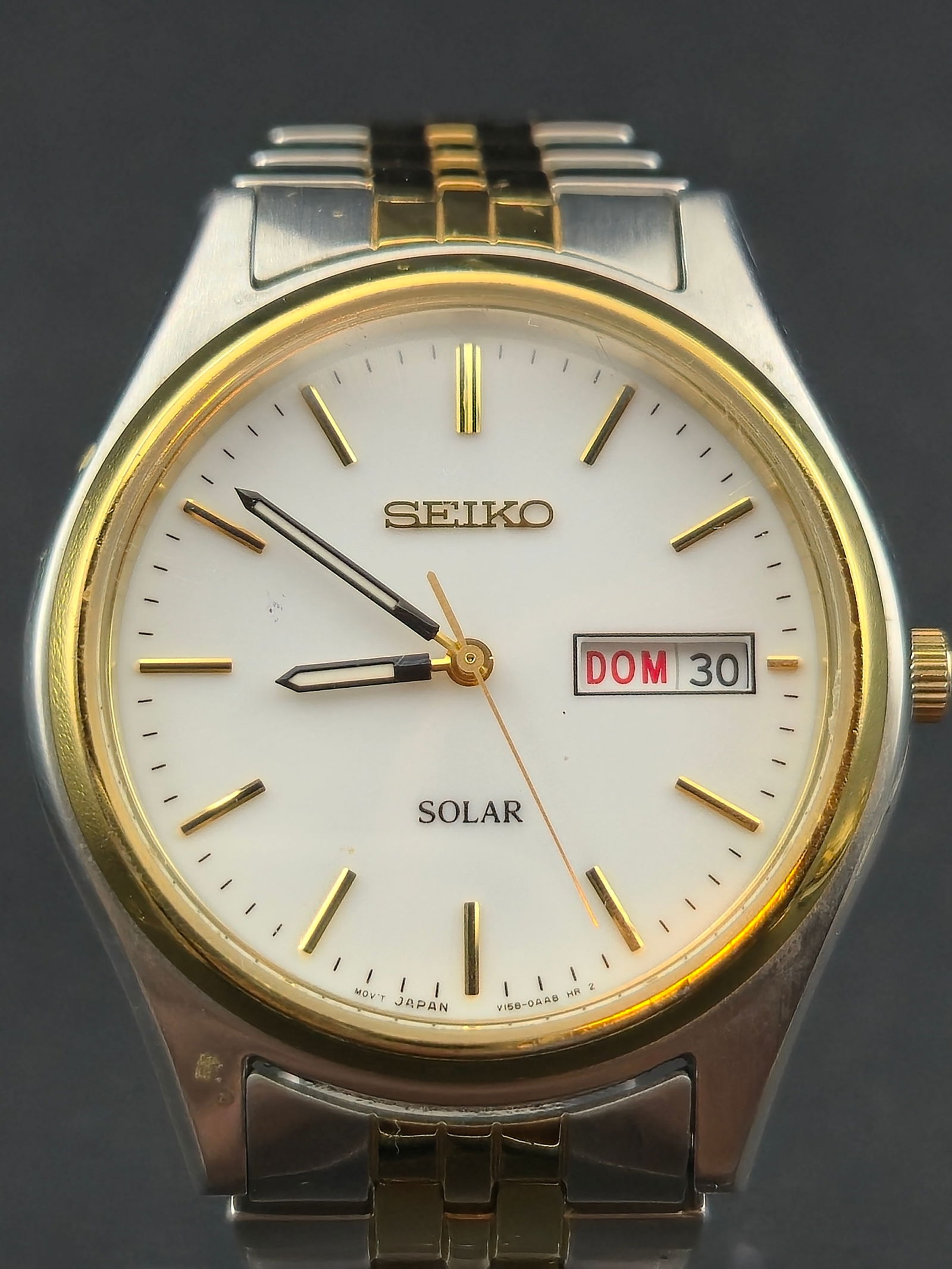 2000s Seiko Solar Day Date Watch Two Tone Stainless Steel 37mm Running: Seiko solar powered quartz wristwatch featuring a white dial with gold tone applied markers and hands. Day and date display at 3 o’clock with bilingual day wheel visible. Watch is running and chargi