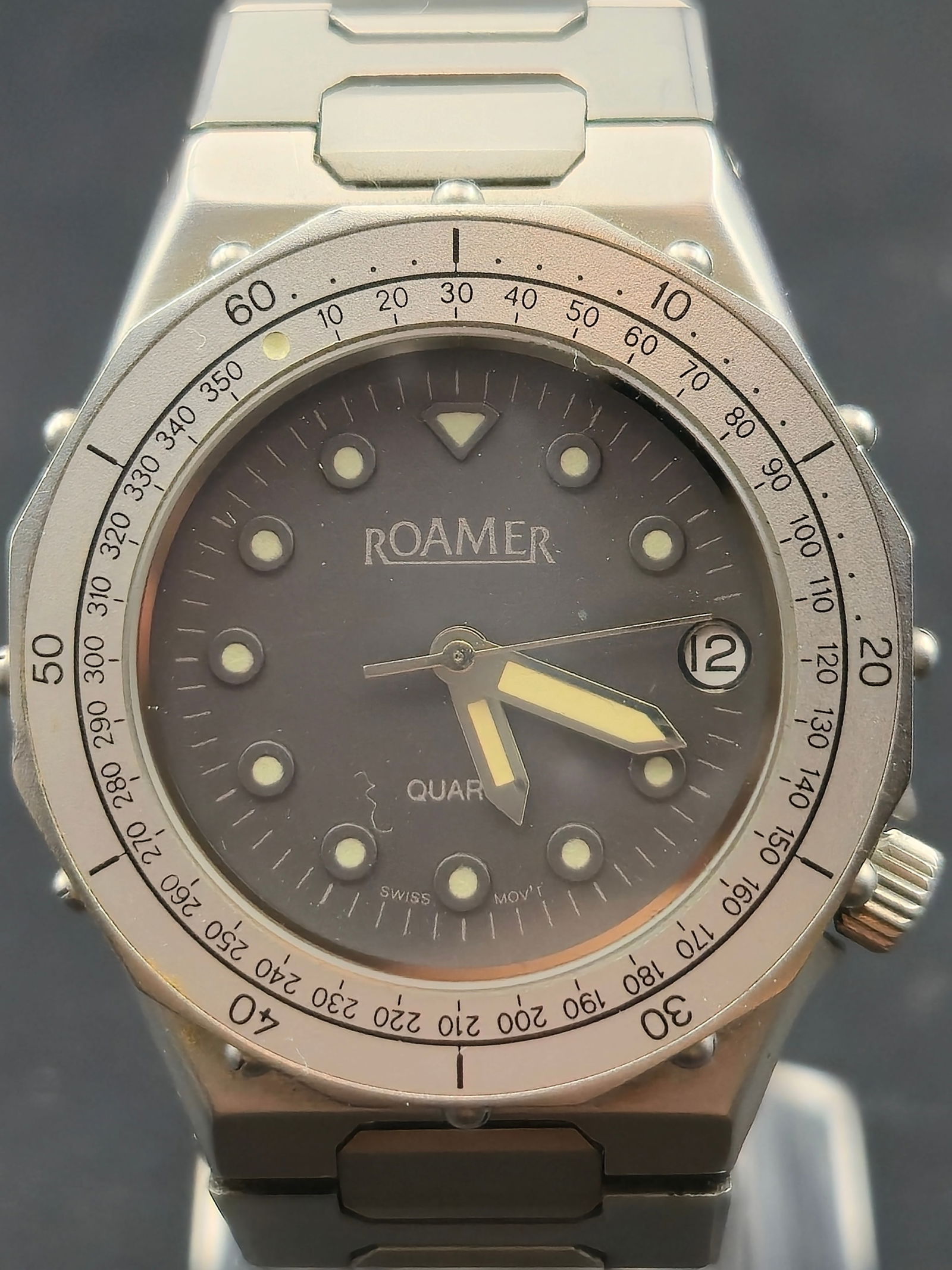 .1990s Roamer Quartz Stainless Steel Date Watch 33mm Swiss Running: Roamer quartz wristwatch with stainless steel case and matching bracelet. Dark dial with luminous round hour markers, triangle marker at 12, and date window at 3 o’clock. Fixed bezel with minute sca