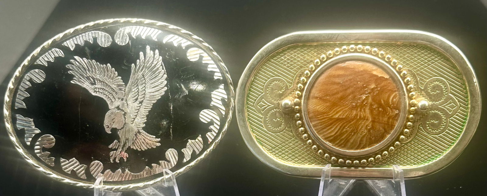 Two Vintage Western Belt Buckles – Eagle & Stone: Pair of vintage Western-style belt buckles. One features a raised eagle on black enamel marked “USA” on the reverse. The second is a gold-tone buckle with a central brown stone accent and text