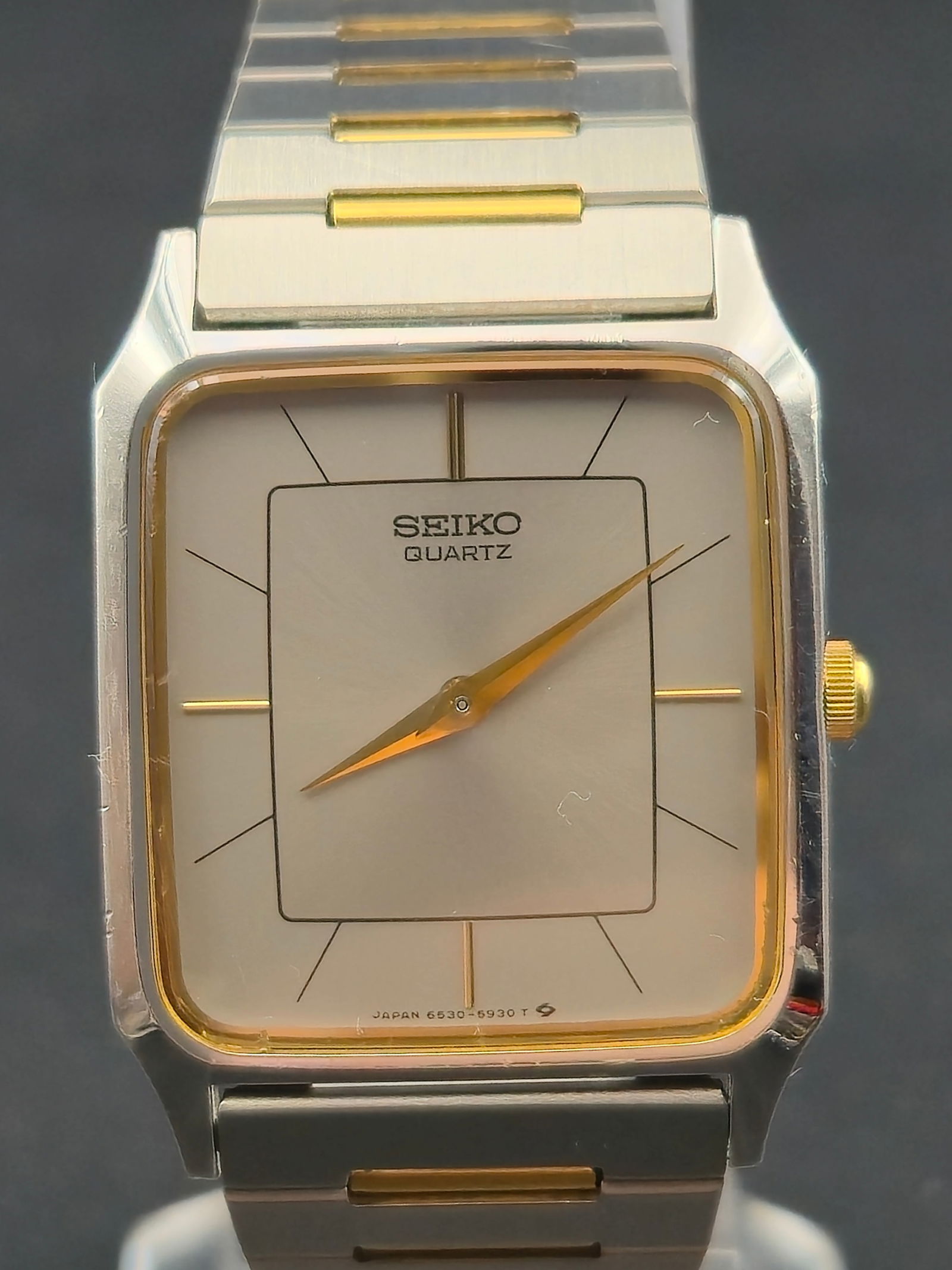 Seiko Quartz Rectangular Two Tone Base Metal Wristwatch 26mm circa 1980s: Seiko quartz rectangular wristwatch featuring a silver dial with minimalist line markers and gold-tone hands. Two-tone rectangular case with gold-tone inner bezel, fitted to a matching two-tone stainl