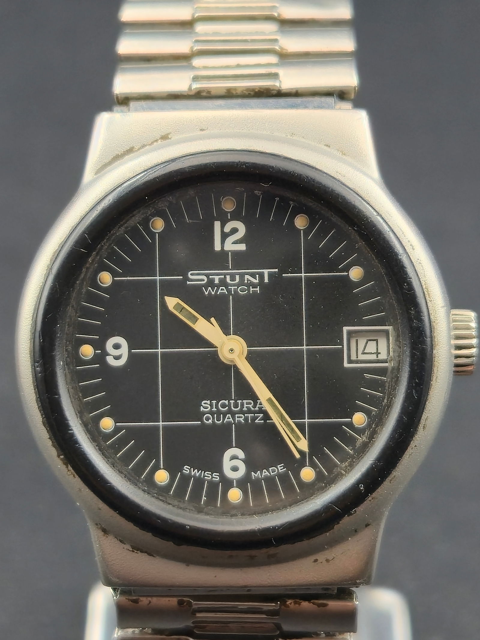 Sicura Stunt Watch Quartz Swiss Made Date Wristwatch 32mm Stainless Steel circa 1980s: Sicura “Stunt Watch” quartz wristwatch featuring a black dial with grid pattern, applied Arabic numerals at 12, 9, and 6, luminous dot hour markers, and a date window at 3 o’clock. Dial is s