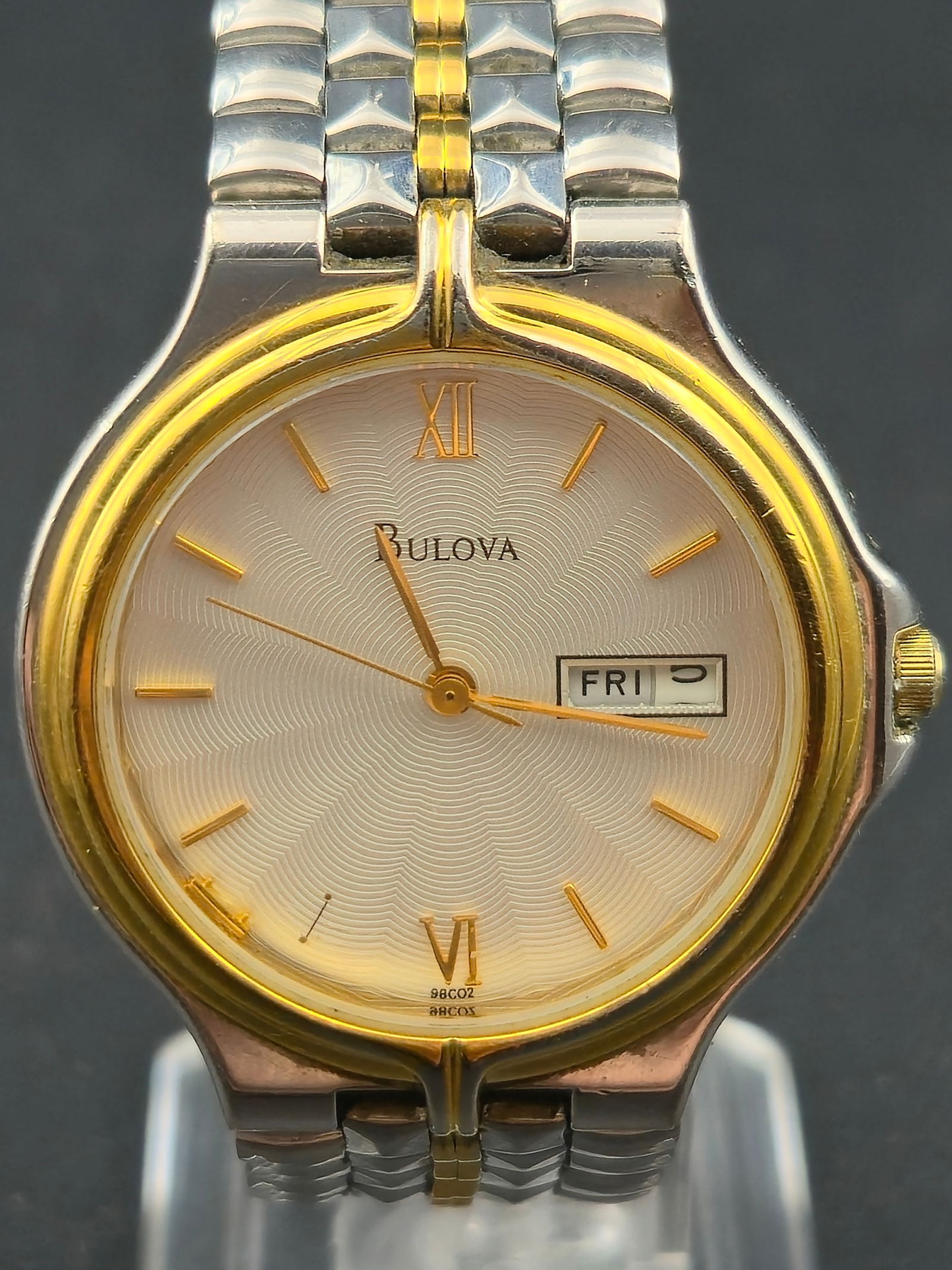 Bulova Quartz Day Date Two Tone Wristwatch Silver Dial 35mm circa 1990s: Bulova quartz wristwatch featuring a silver guilloché-style dial with concentric wave pattern, applied gold-tone baton hour markers, and Roman numeral XII and VI. Day-date display at 3 o’clock. Two