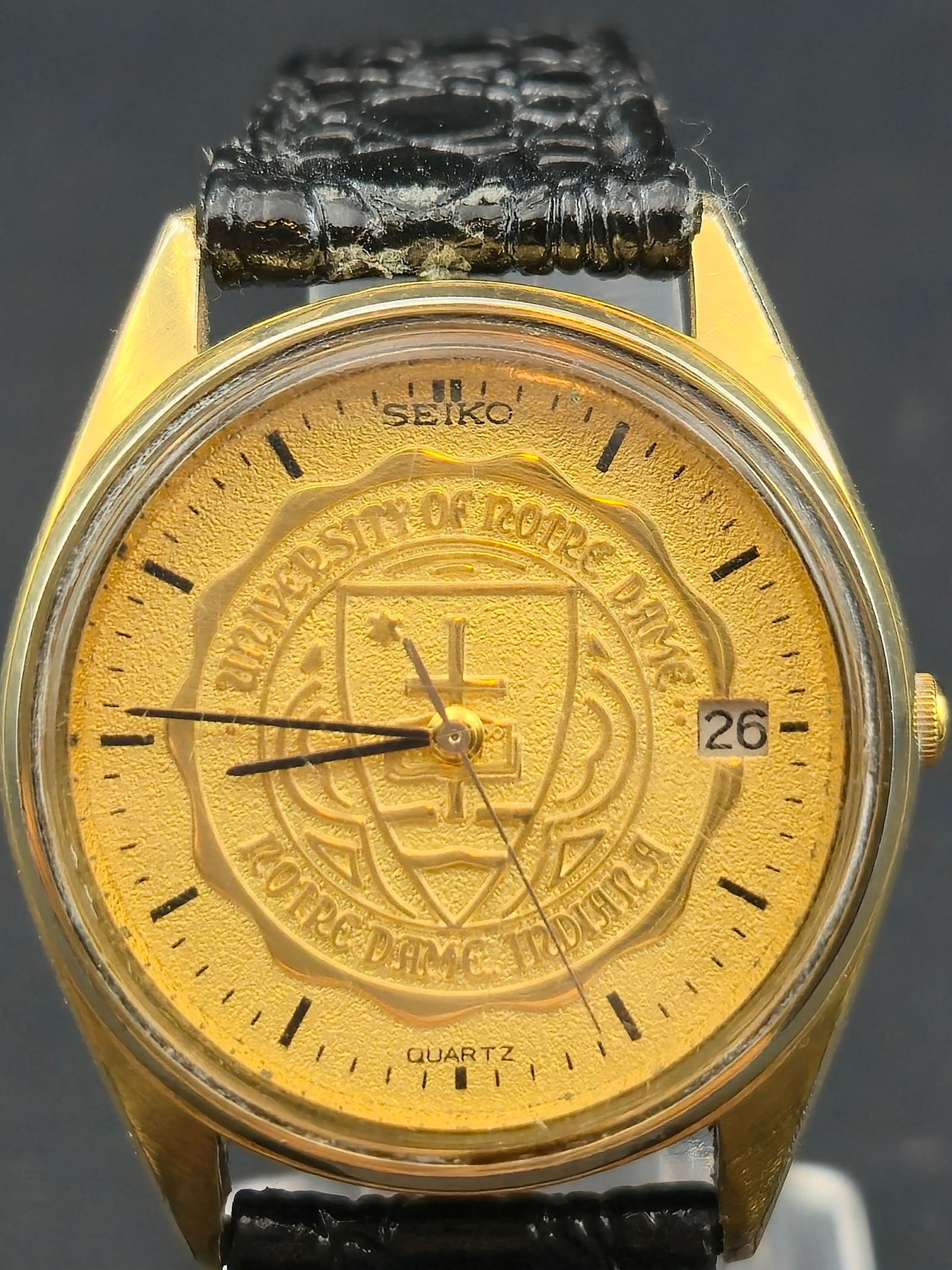 1990s Seiko Quartz 34mm Gold Tone University of Notre Dame Dial Date Watch: Vintage Seiko quartz wristwatch featuring a gold embossed collegiate seal dial reading “University of Notre Dame” with “Notre Dame Indiana” along the lower arc. The raised central ND c