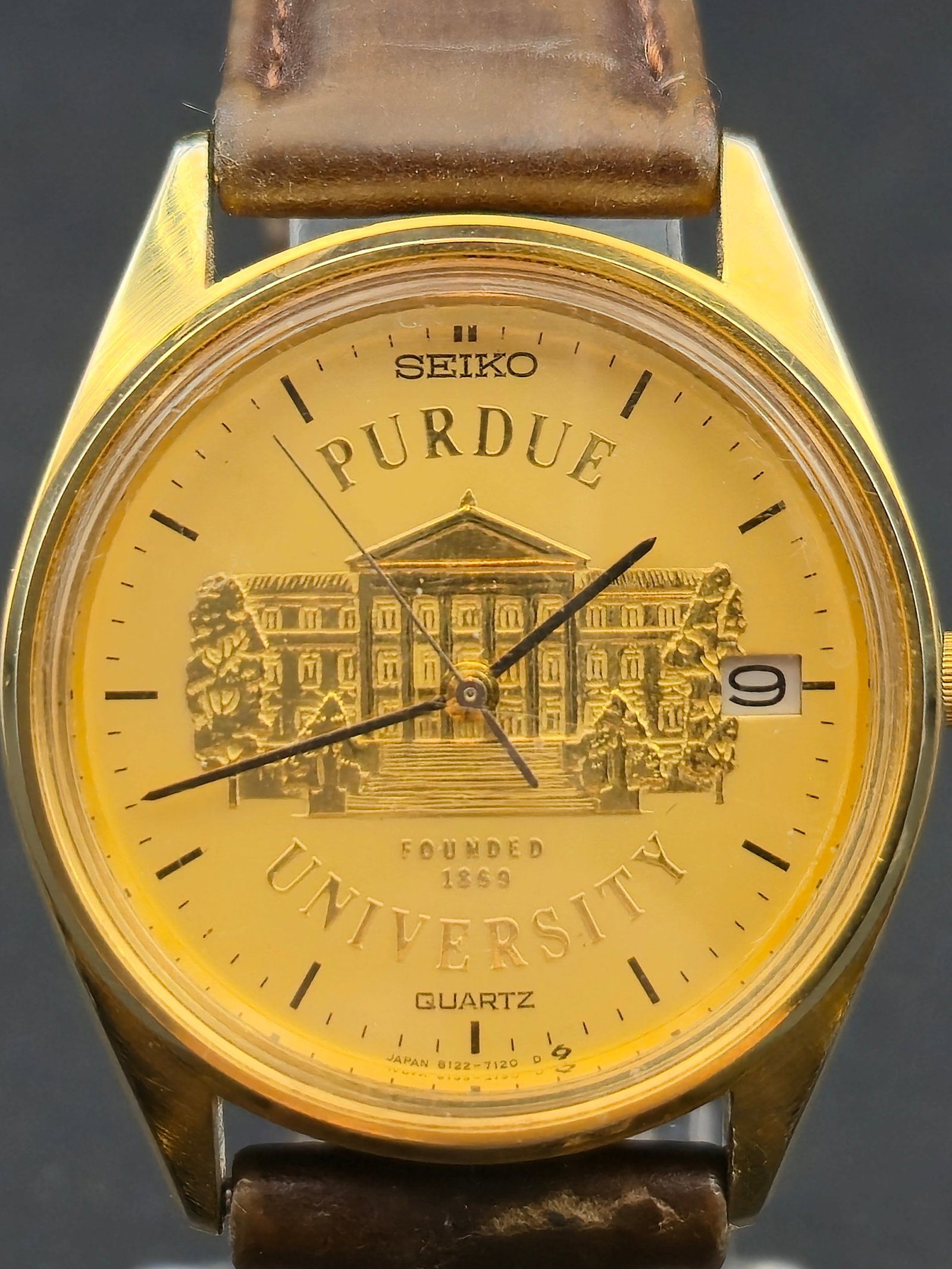 1983 Seiko 8122-7120 34mm Purdue University Quartz Base Metal Watch New Battery: 1983 Seiko 8122-7120 Purdue University quartz wristwatch with gold tone dial featuring campus building motif and “Founded 1869.” Black baton hour markers with date at 3 o’clock. Base metal c