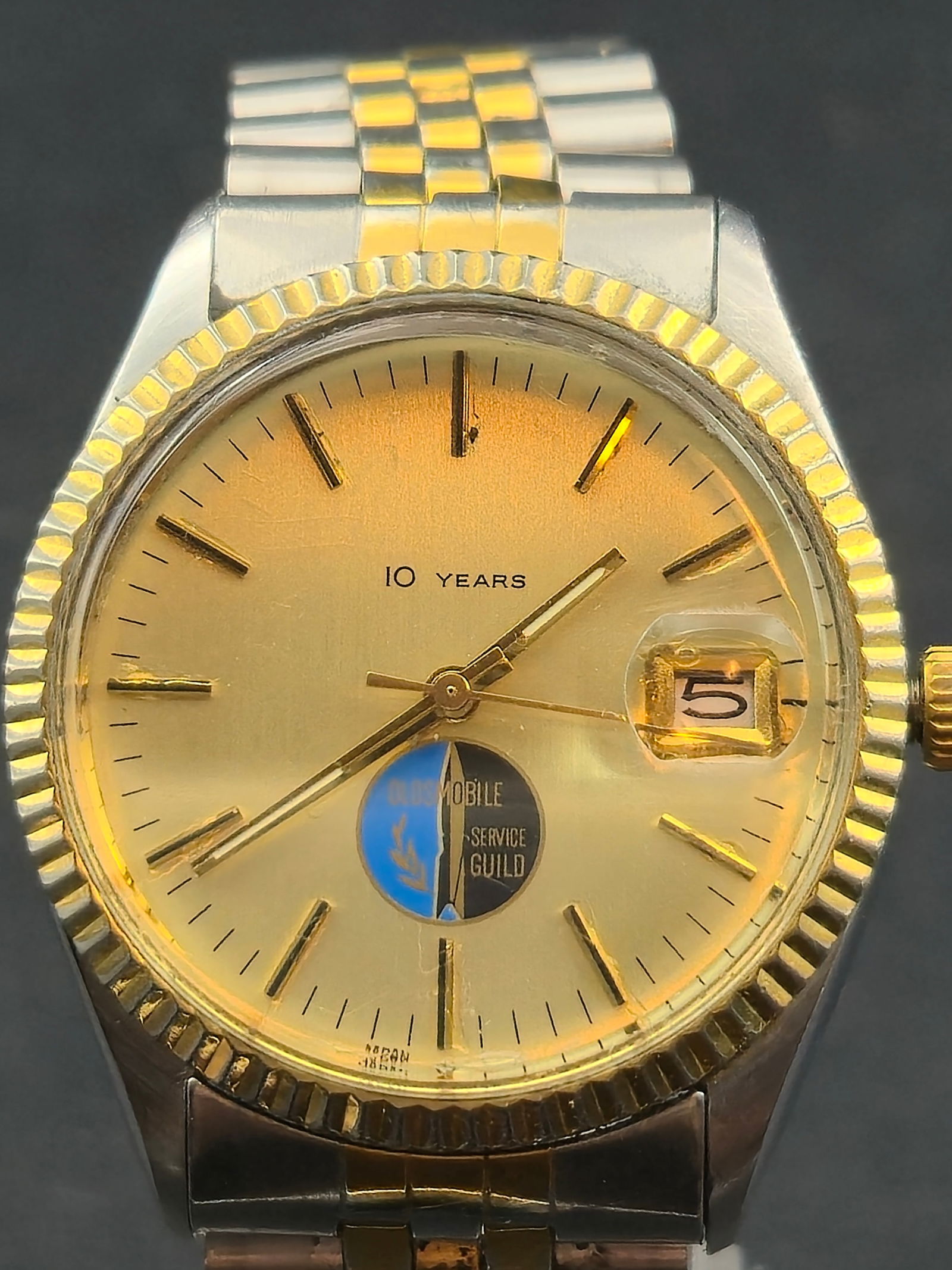 Pulsar 10 Years Oldsmobile Service Guild Watch Gold Tone 10ATM Date 35mm Quartz: Vintage Pulsar quartz wristwatch featuring a gold tone dial with applied markers and a distinctive Oldsmobile Service Guild 10 Years commemorative medallion at 6 o’clock. The watch has a fluted beze