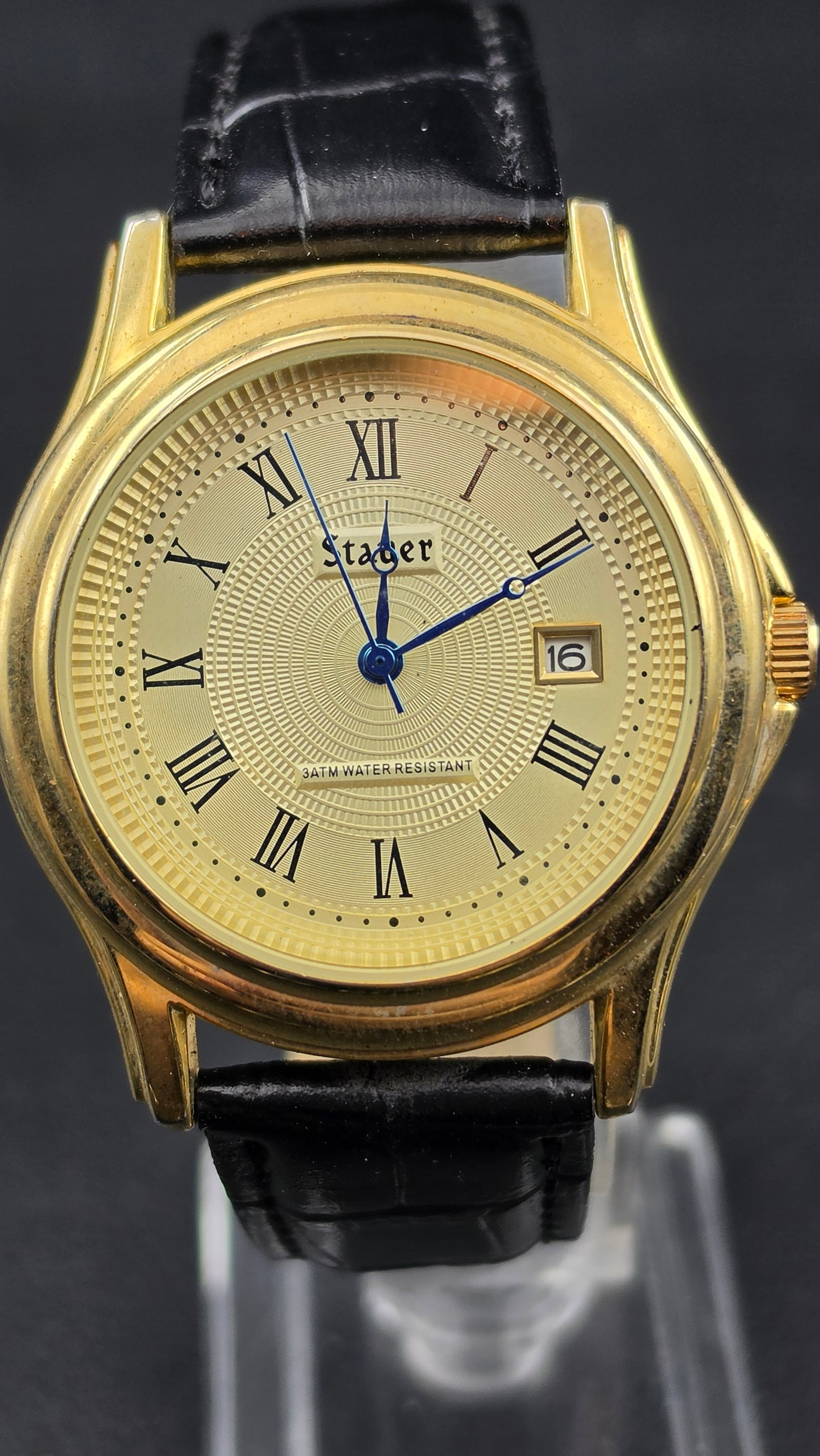 Stauer Gold Tone Roman Dial Date Watch Blue Hands 3ATM Leather Strap 1990s New Battery Running: Stauer dress watch featuring a guilloché-style textured gold dial with black Roman numerals and blued steel-style hands. Date window at 3 o’clock and “3ATM Water Resistant” printed on dial.